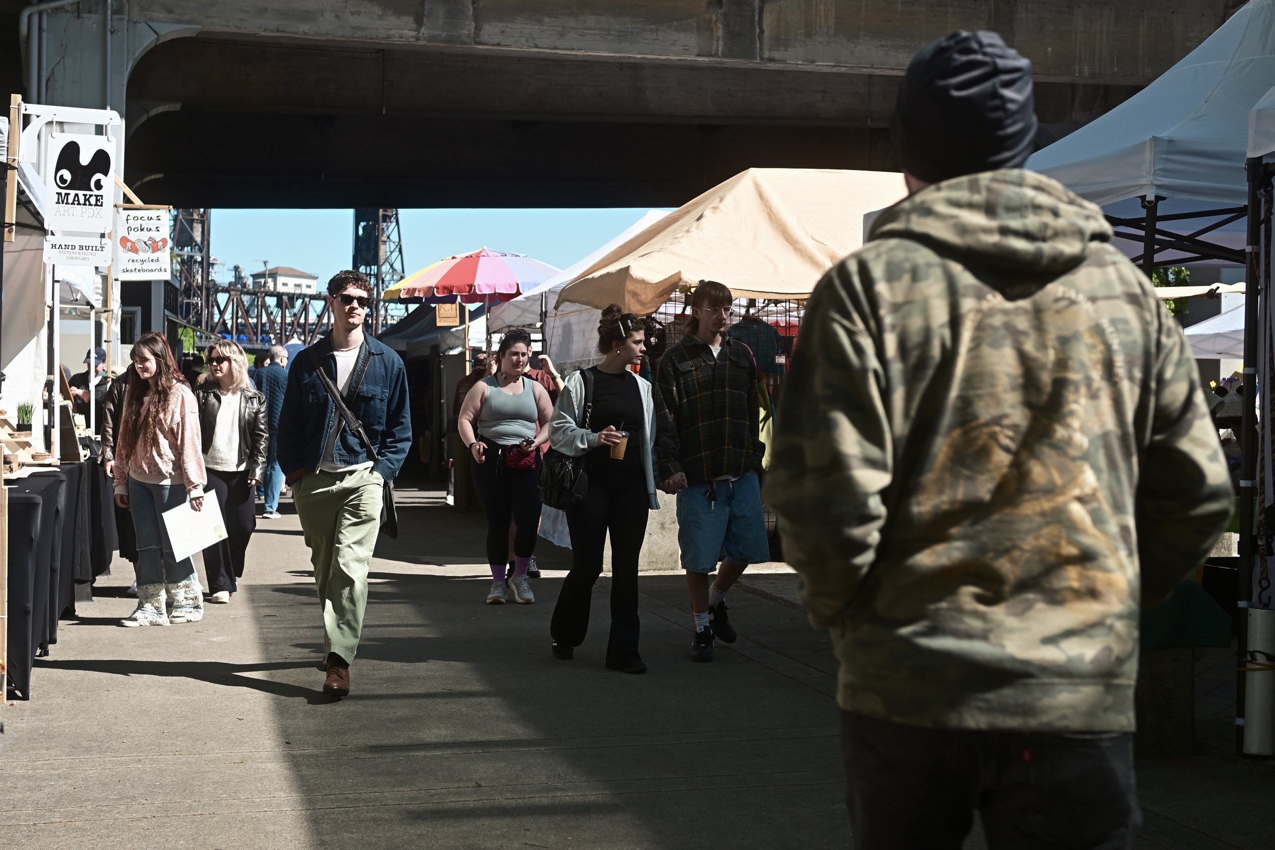 People walking at an outdoor market under a bridge, with stalls, umbrellas, and vendor signs visible.