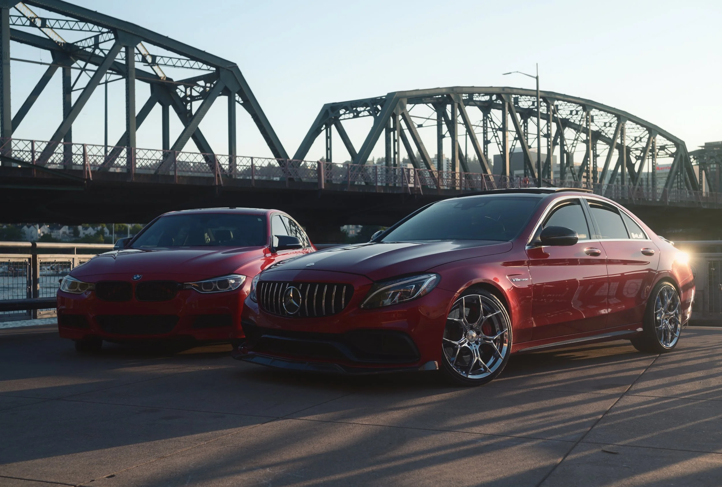 Two red luxury cars, a BMW and a Mercedes-Benz, parked near a bridge over a body of water during sunset.