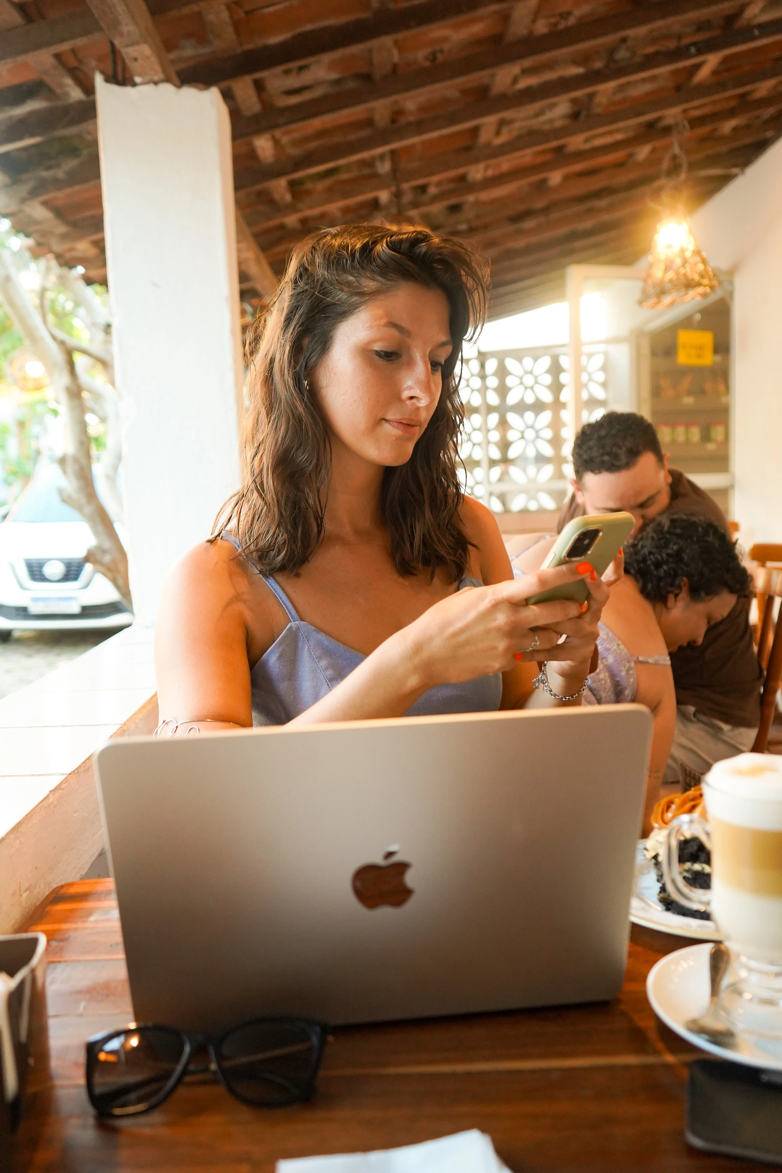 Una mujer con el pelo castaño y ondulado que mira su móvil, sentada en una mesa de madera en un restaurante o una cafetería. Delante de ella hay un portátil MacBook plateado. Hay otras personas sentadas cerca. Hay bebidas sobre la mesa y unas gafas d