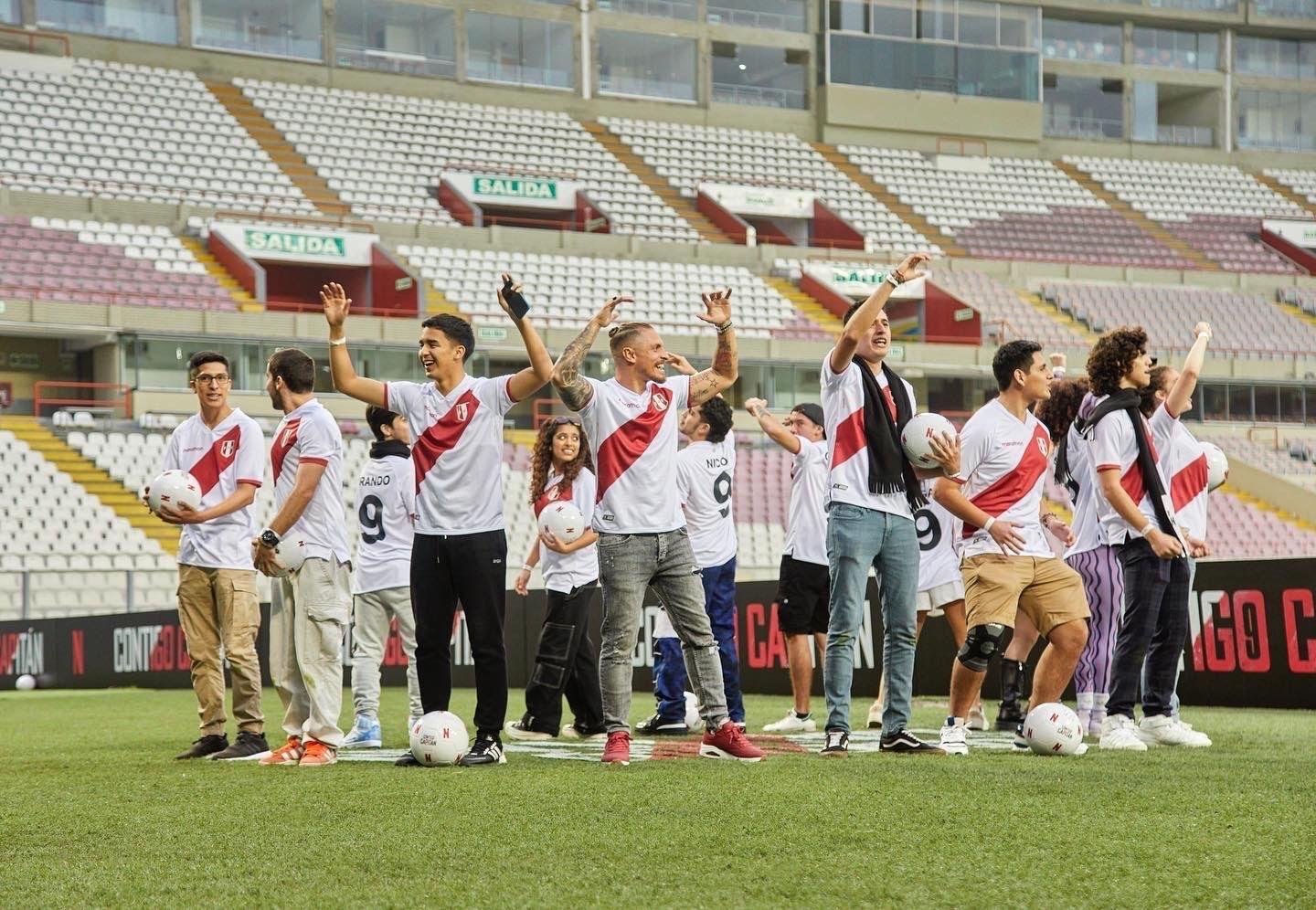 Un grupo de futbolistas y miembros del cuerpo técnico con camisetas blancas con una franja diagonal roja, de pie en el campo de fútbol de un estadio, saludando con la mano y sonriendo, con balones de fútbol en las manos, y con las gradas vacías al fo