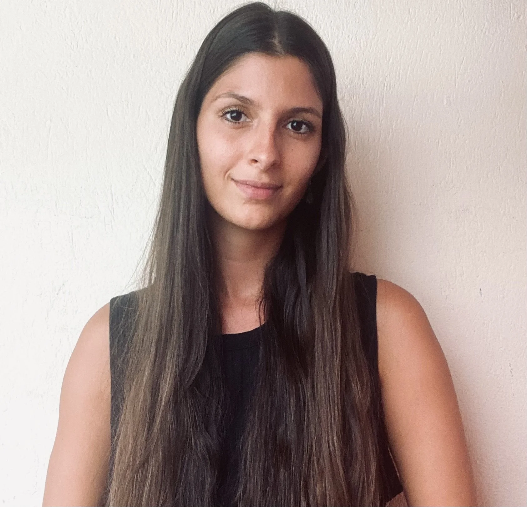Retrato de una mujer con el pelo largo y oscuro que lleva un top negro sin mangas, de pie frente a una pared lisa de color claro.
