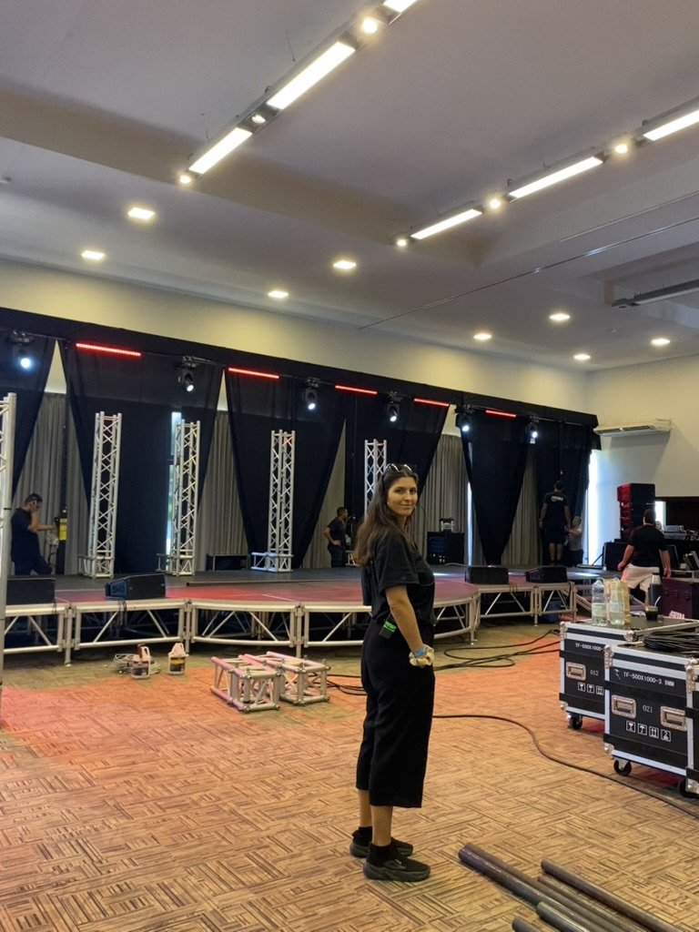 Una mujer con el pelo largo y castaño, vestida de negro, de pie en un espacio cubierto destinado a eventos, con un escenario, equipo de iluminación y varias personas más preparando el escenario.