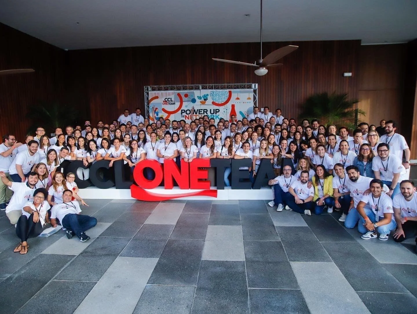 Un numeroso grupo de personas en un evento, de pie detrás de unas grandes letras en 3D con nombre de una convención, con algunas personas sentadas o arrodilladas delante y otras de pie al fondo, en una sala con paneles de madera en las paredes y plan