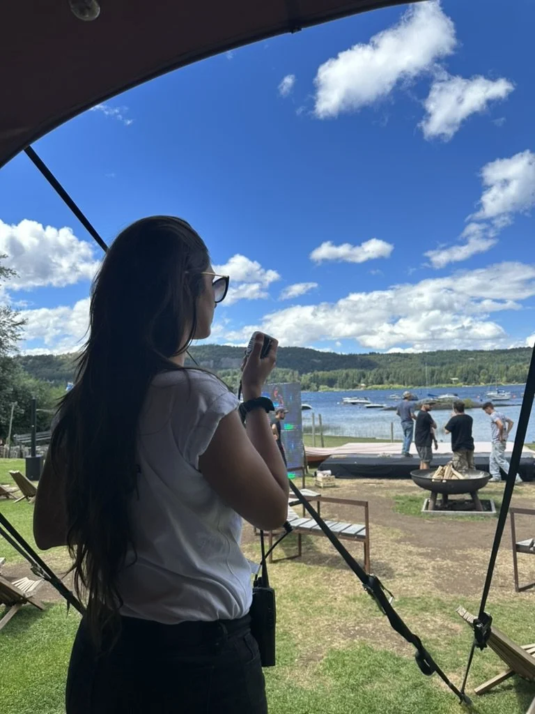 Mujer con el pelo largo y gafas, de pie bajo un toldo, sosteniendo un teléfono cerca de un lago con barcas, algunas personas alrededor de una hoguera y un cielo azul con nubes.