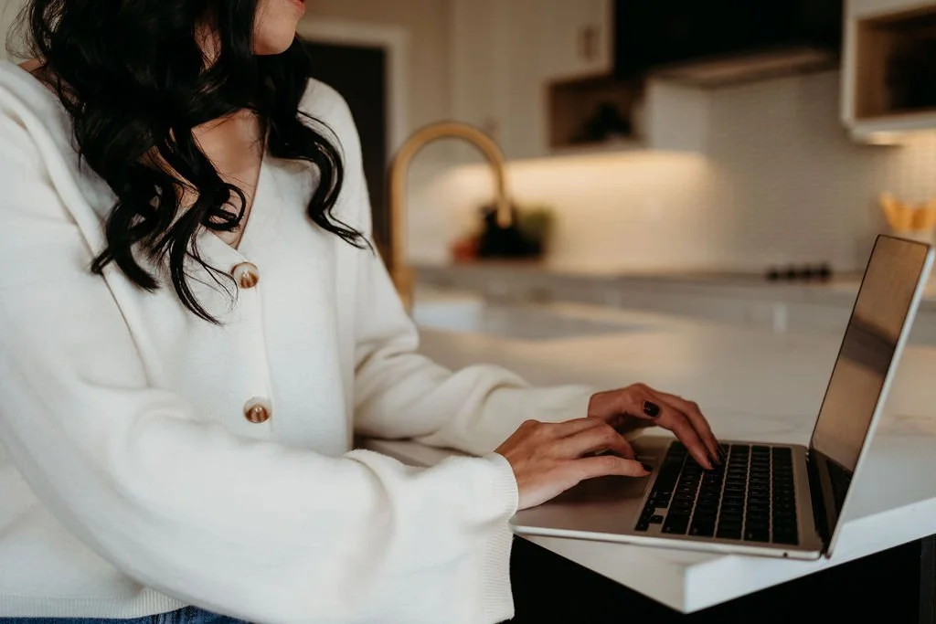 Person wearing a white sweater typing on a laptop in a kitchen setting.