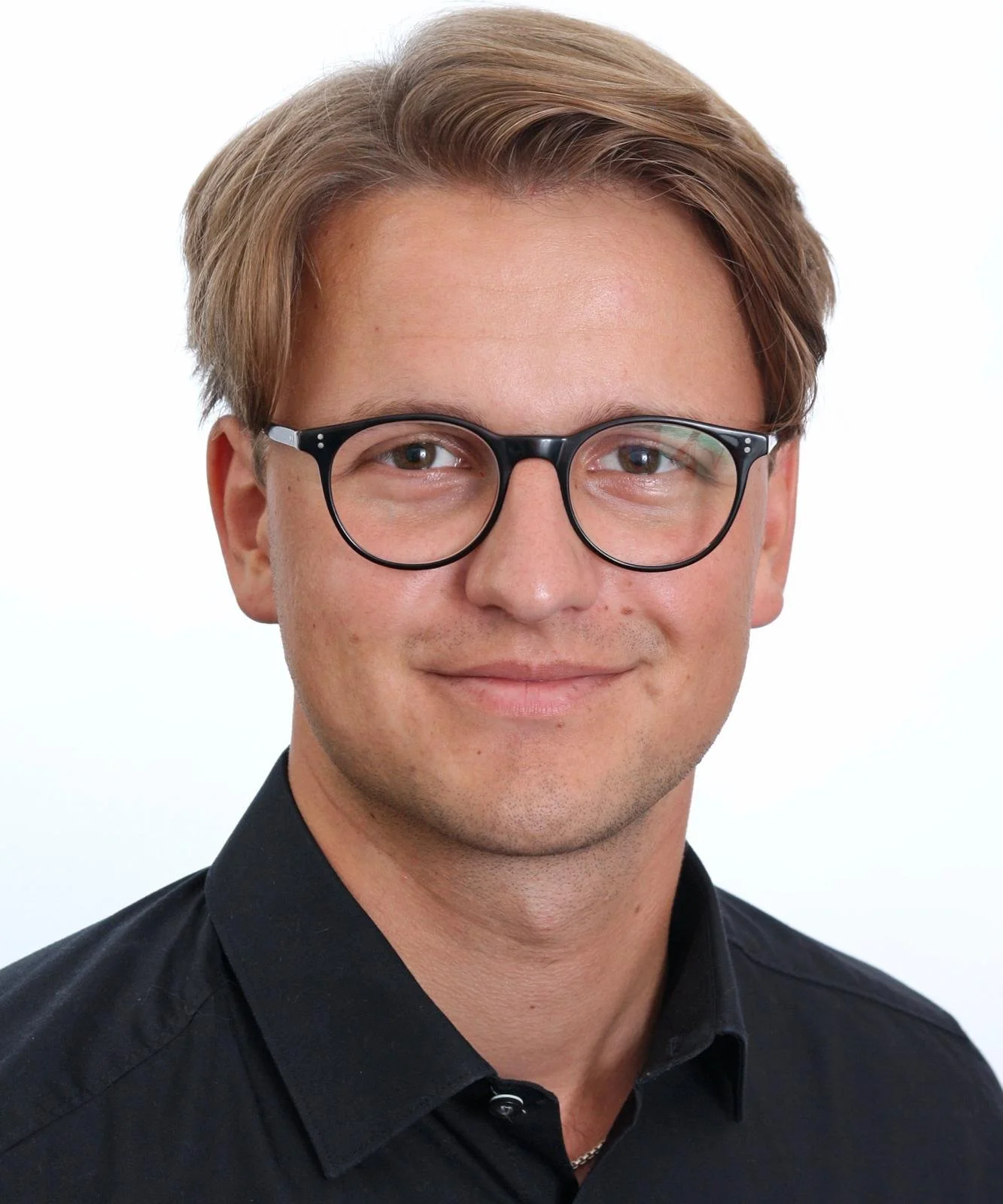 Headshot of a young man with light skin, light brown hair, glasses, and a black shirt, smiling against a white background.