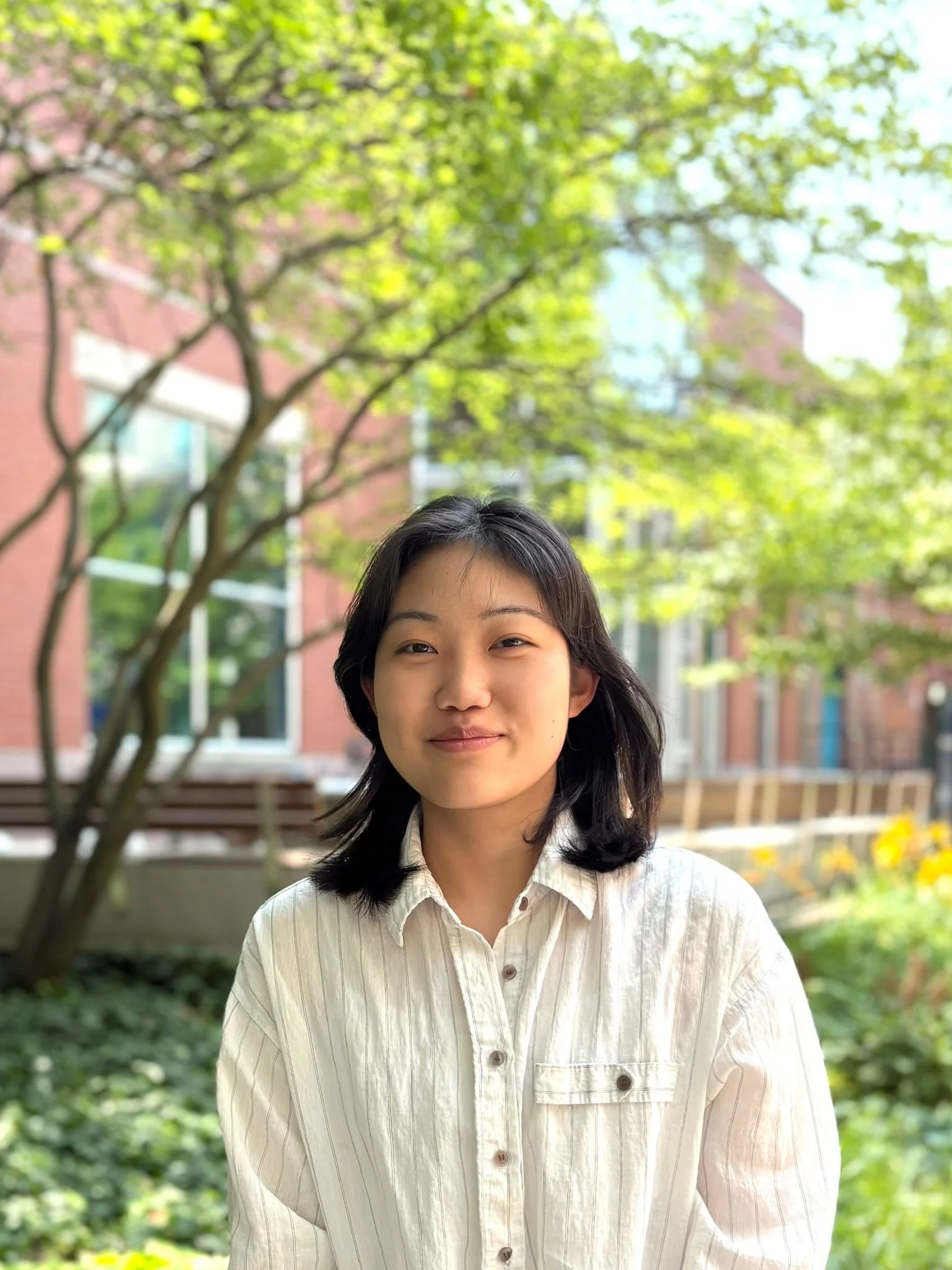 Young woman smiling outdoors with trees and a building in the background.