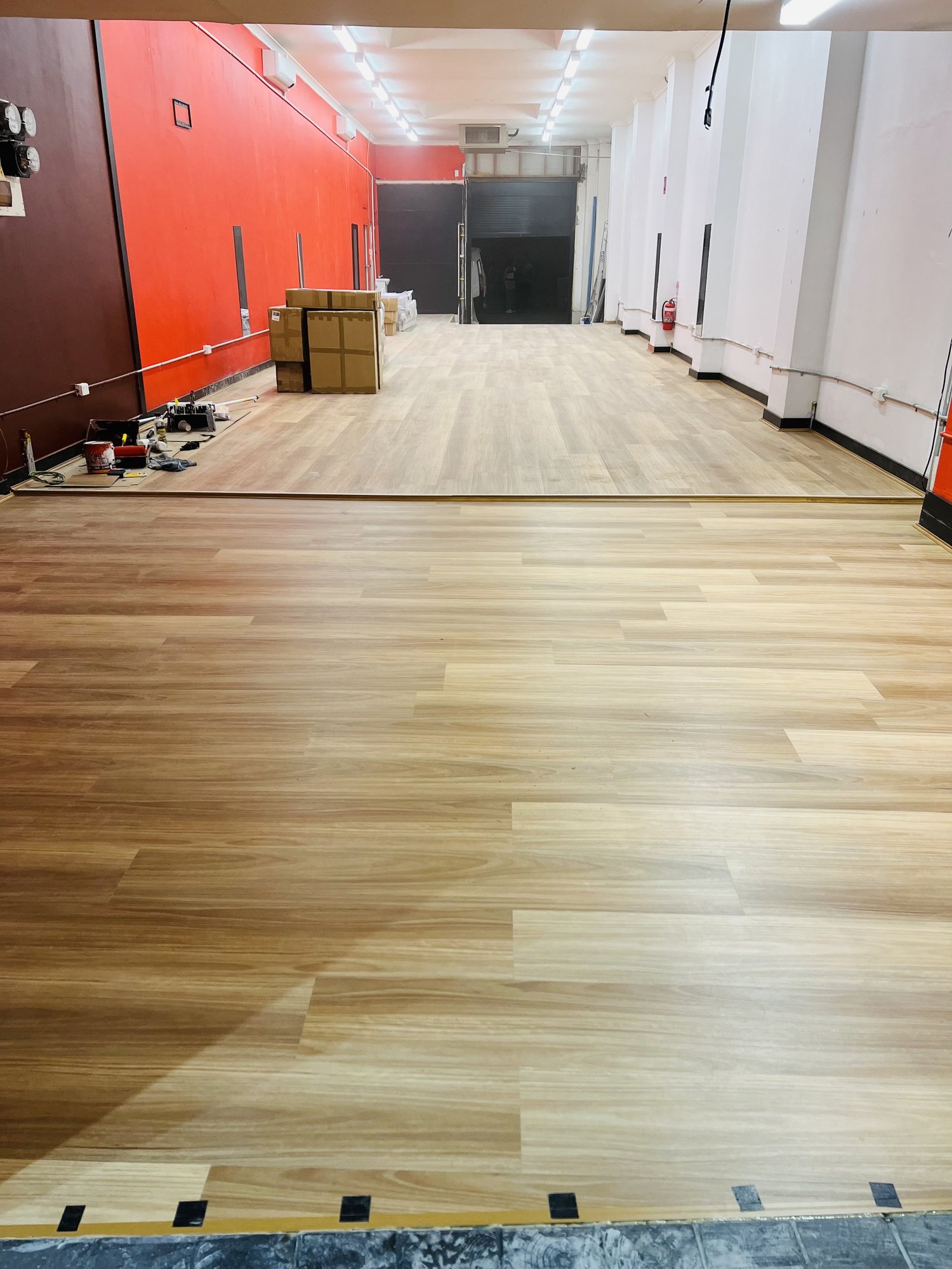 An indoor space with partially installed wooden flooring, construction tools, and supplies. The room has a red accent wall, some white walls, and a black rolling door in the background.