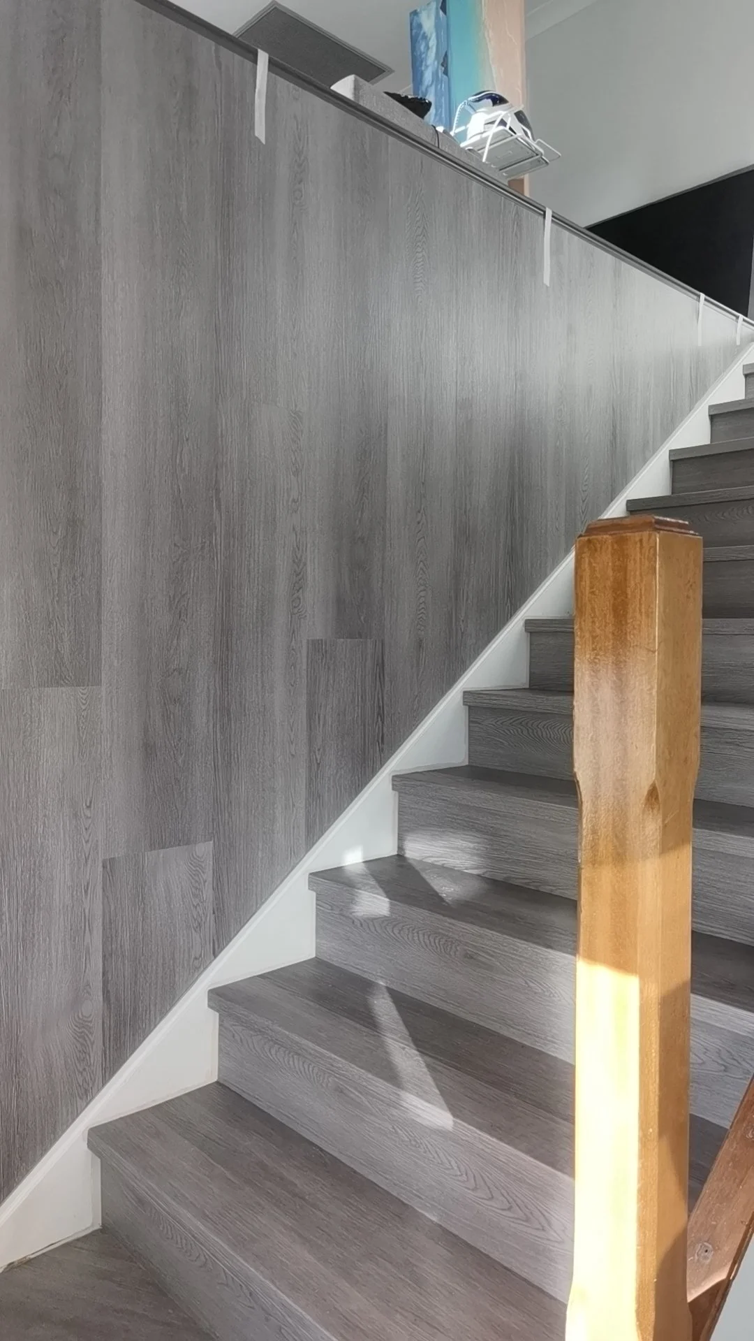 Wooden staircase with gray steps and a wooden banister, next to a wall with wooden paneling, under natural light.