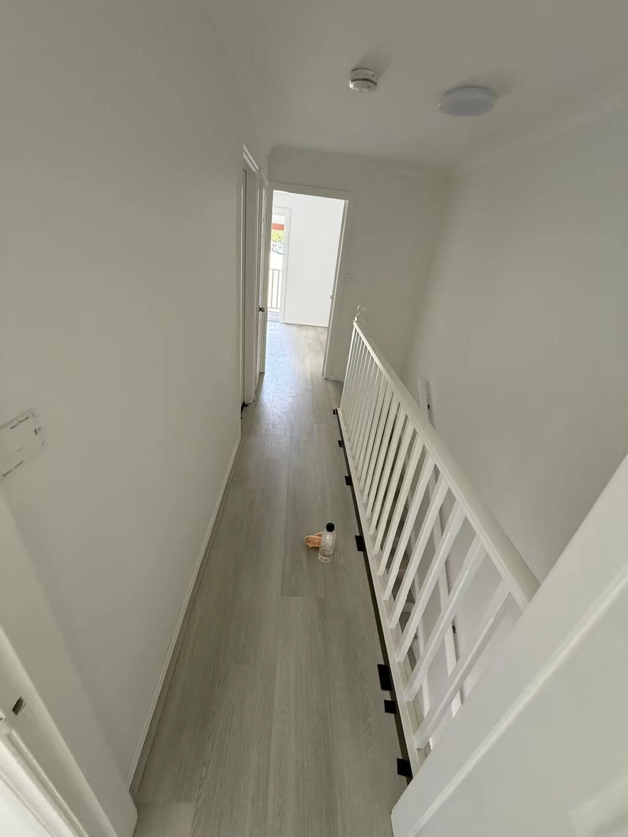 Empty hallway with white walls, a water bottle and cloth on the floor, a white railing on the right, and an open door leading outside in the distance.