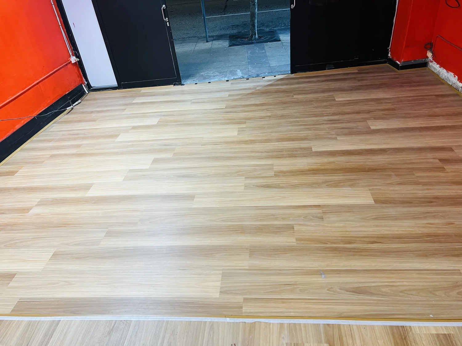 New light wood laminate flooring in a room with red and black walls, with an open door leading outside to a sidewalk and tree.