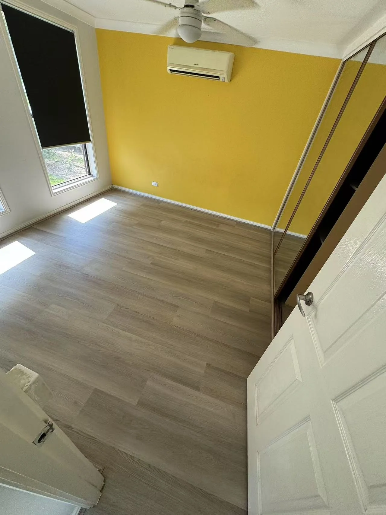 Empty bedroom with light wood flooring, yellow accent wall, white door partially open, built-in closet with sliding mirrors, large window with black blinds, ceiling fan, and wall-mounted air conditioning unit.