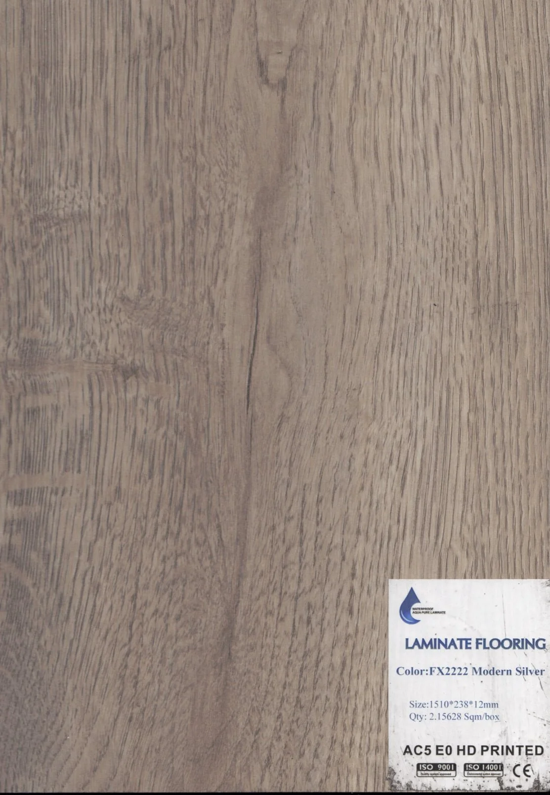 A close-up photo of a wooden laminate flooring sample with a label indicating the color 'Modern Silver' and specifications.