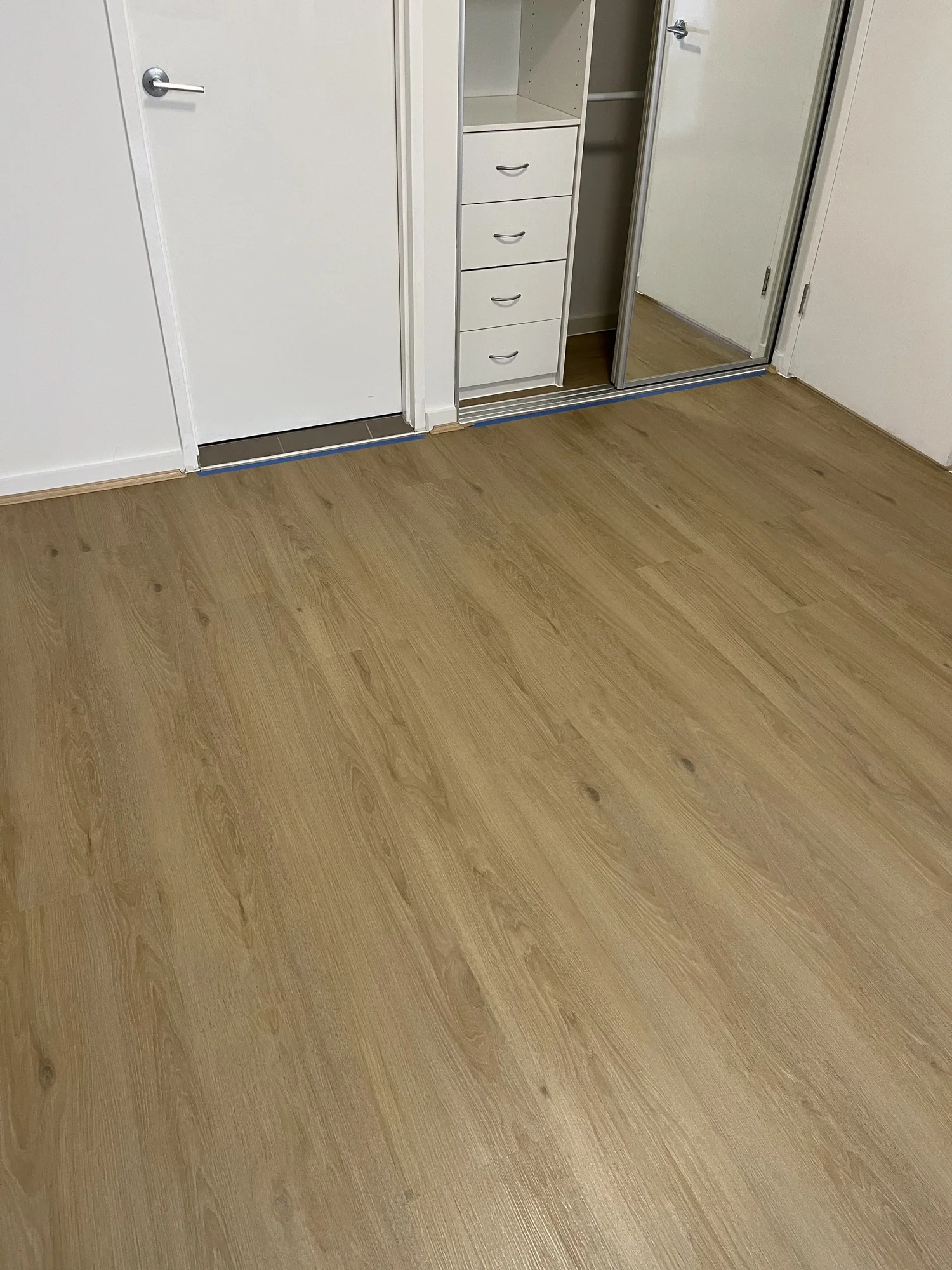 A room with light-colored wooden flooring, a white door, a closet with a sliding mirror door, and a small white dresser with four drawers.