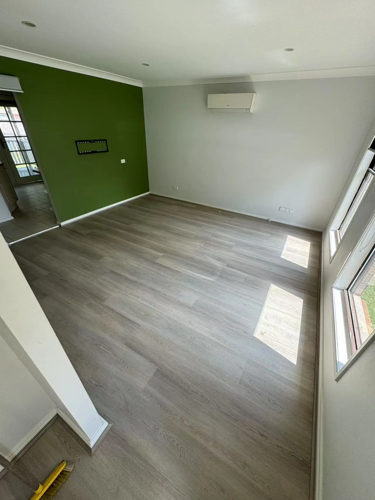 Empty living room with wooden flooring, a green accent wall, large windows on the right allowing sunlight, and a white wall with an air conditioning unit.