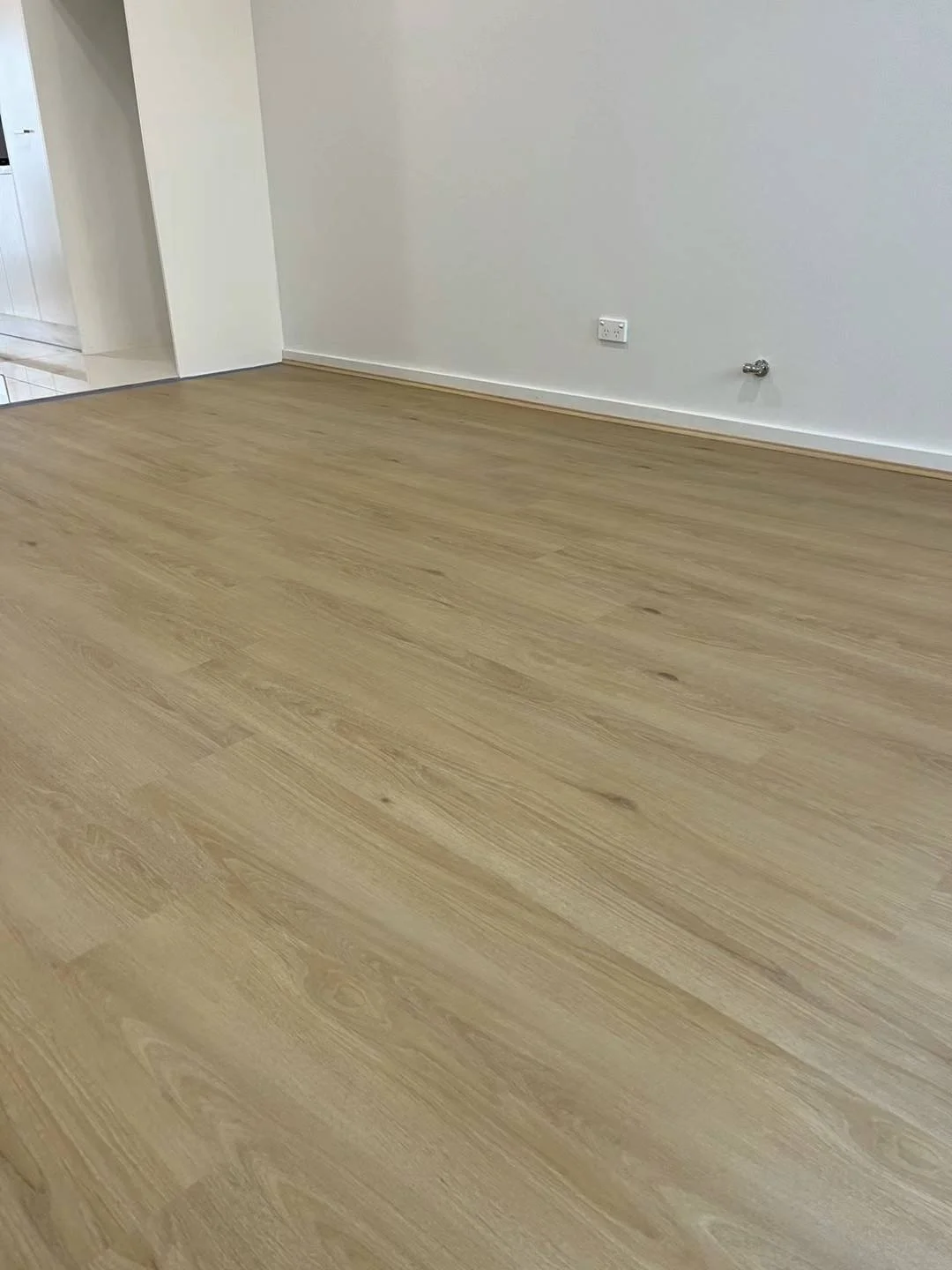 Room with light wooden laminate flooring, white walls, an electrical outlet, and a small pipe on the wall.