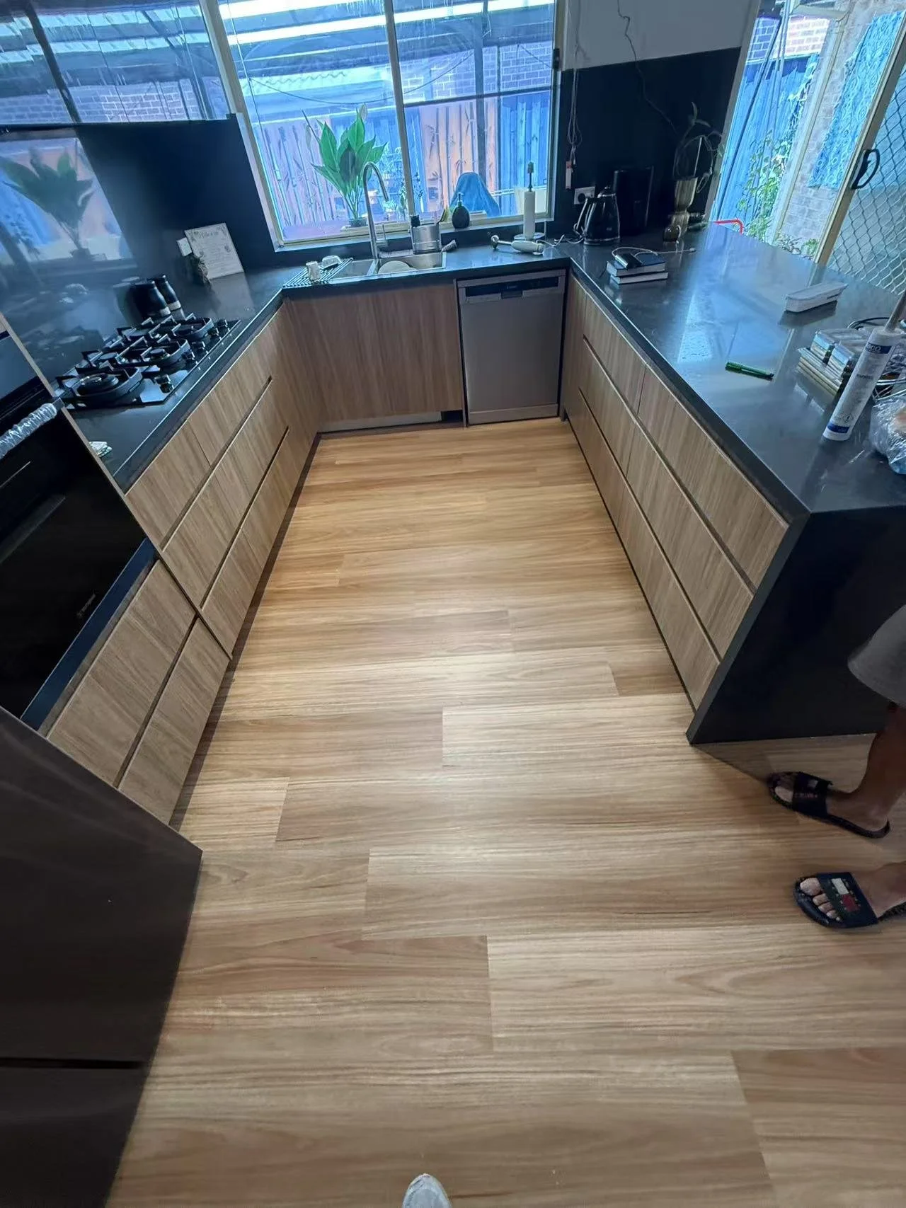 A modern kitchen with black countertops, wooden cabinets, a window with plants, and a view of a backyard with a fence. There is a sink, appliances, and various items on the counters.