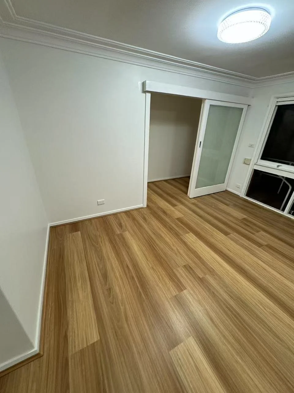 Empty room with white walls, a wooden floor, and sliding glass door leading to a balcony or another room. Ceiling has a modern circular light fixture.