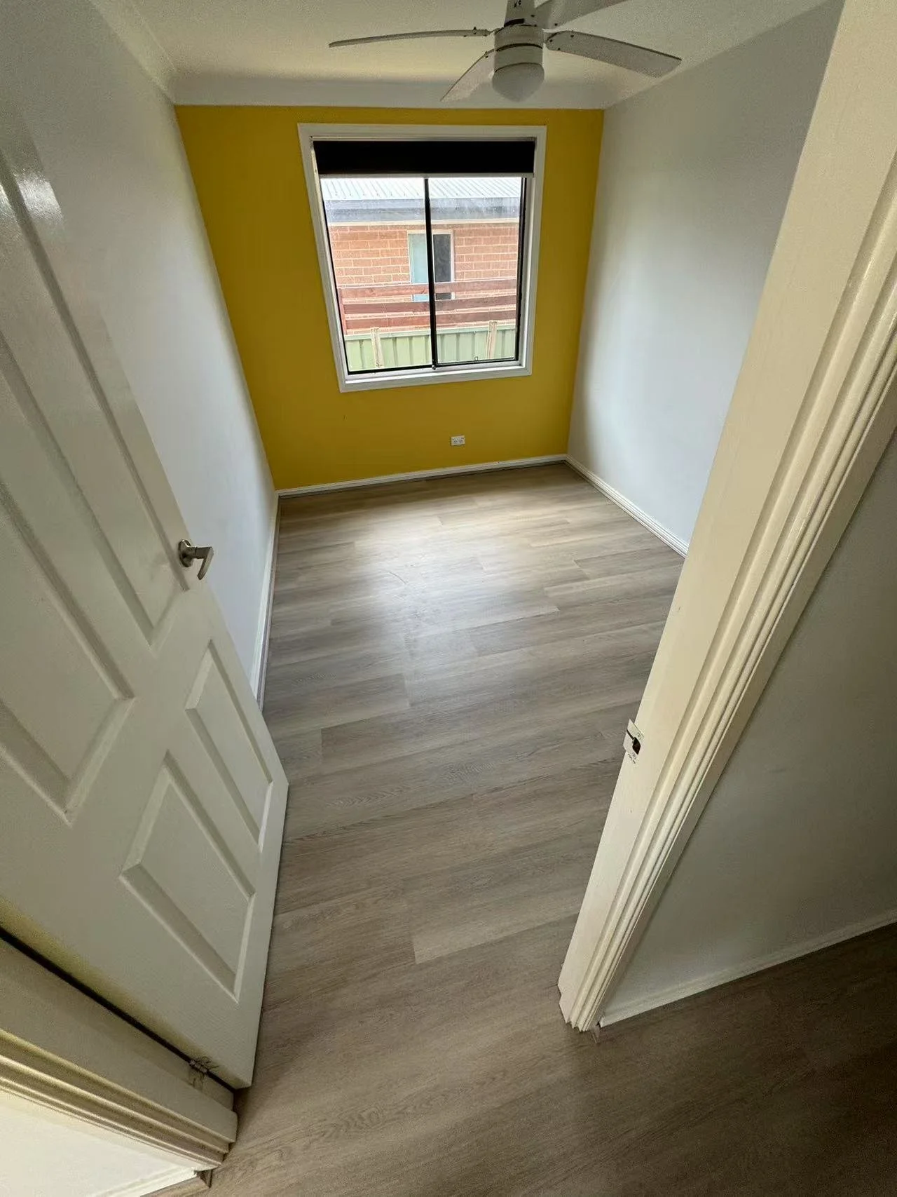 Empty small bedroom with yellow accent wall, hardwood floor, window with black blind, white ceiling fan, and white doors.
