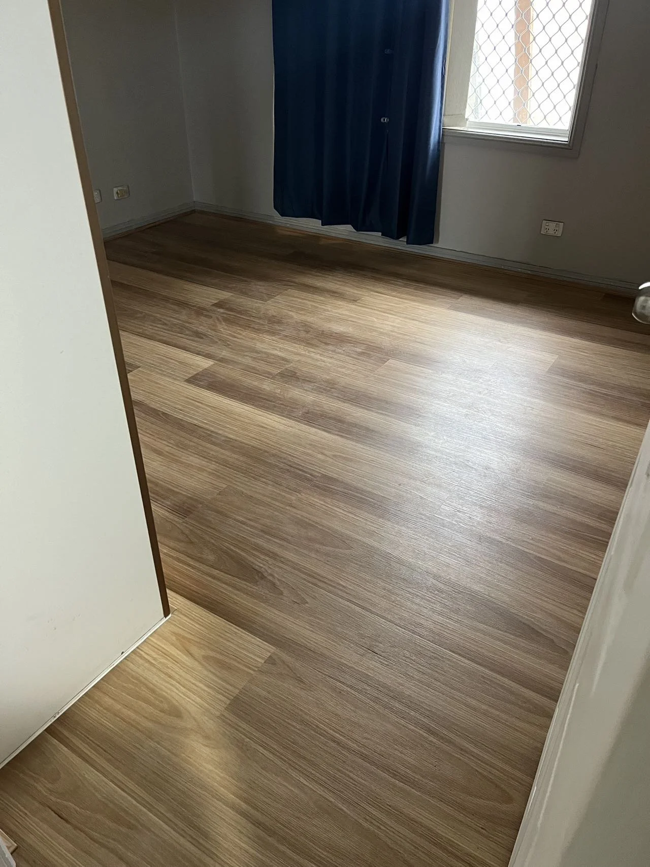 Empty room with wooden flooring, a window with a security screen, and a dark blue curtain.