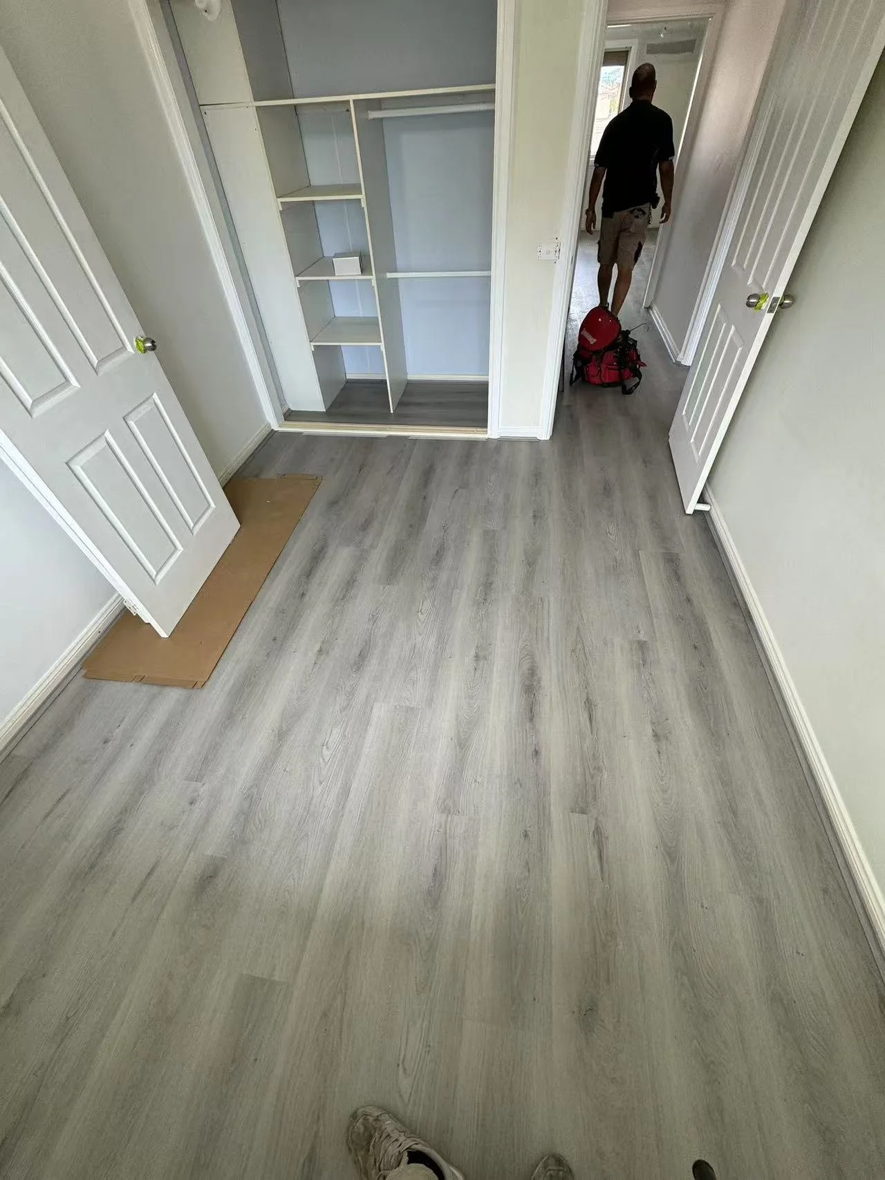 Empty room with light-colored wooden flooring, open closet with shelves, and two open white doors. A person is walking away in the hallway, carrying a red bag.