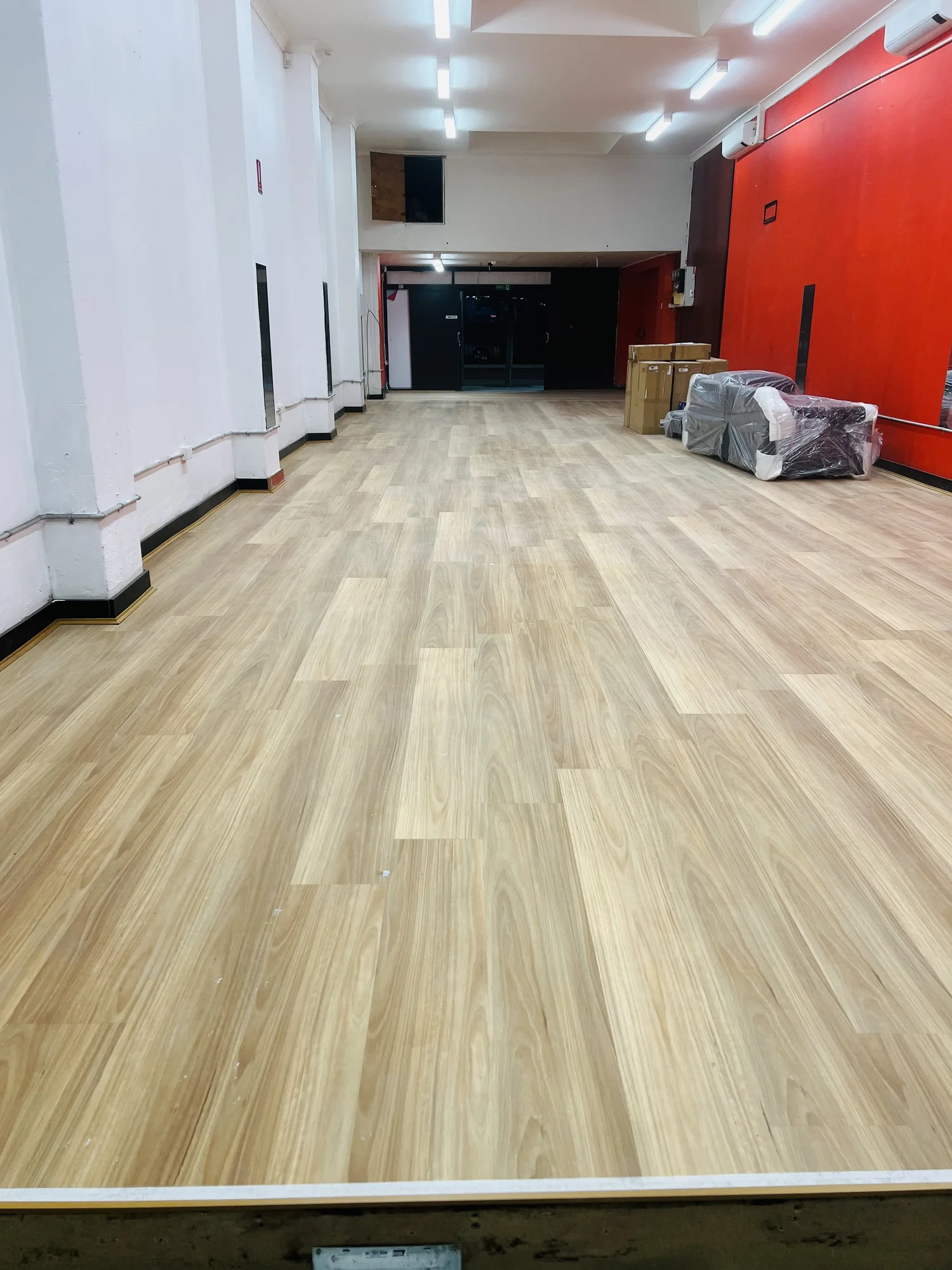 A room with new light wood flooring, white and red walls, and fluorescent lighting, with some furniture wrapped in plastic and cardboard boxes.
