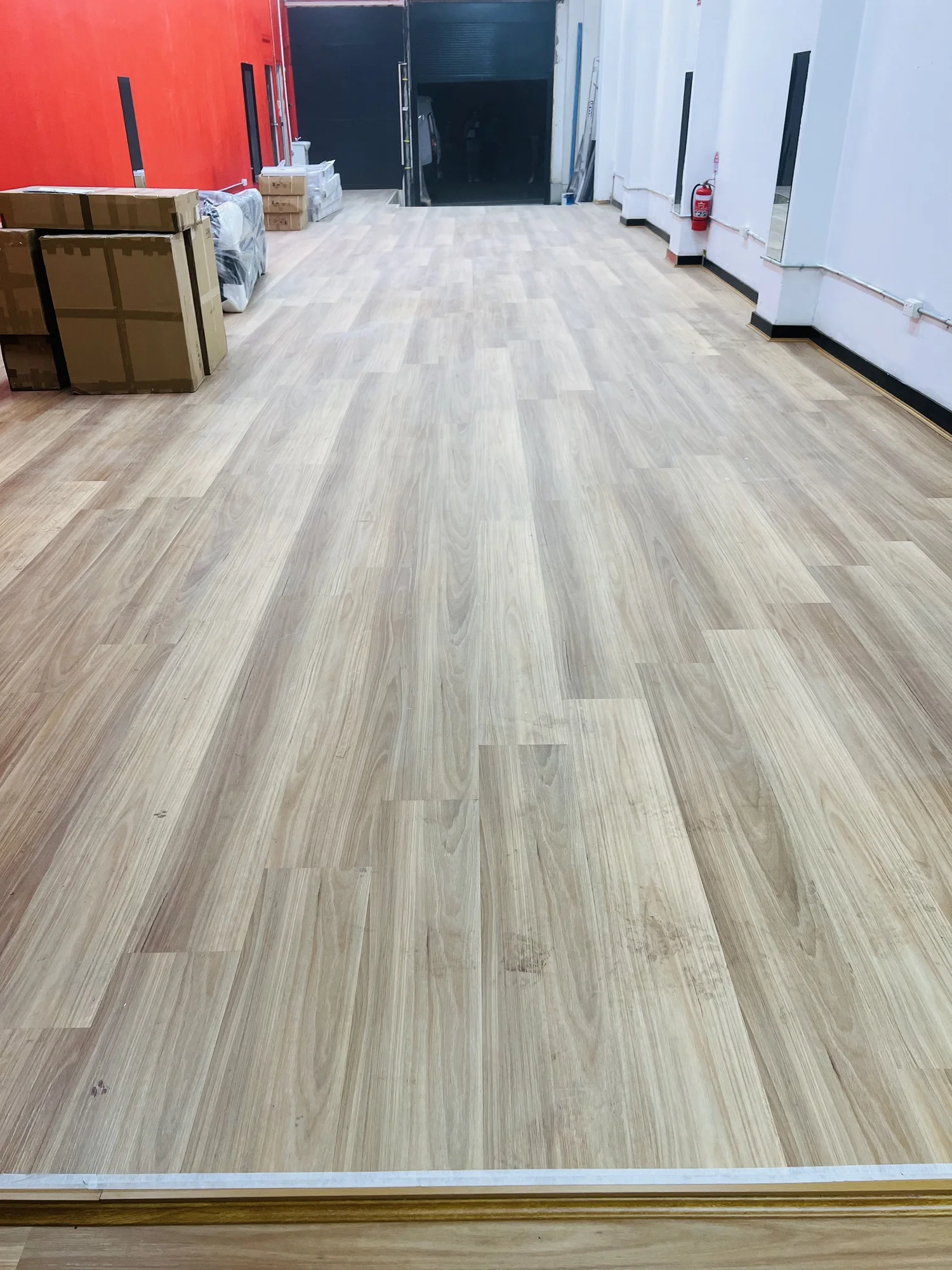 Interior space with new light-colored wooden flooring, red accent wall on the left, white walls on the right, cardboard boxes on the left, and a fire extinguisher mounted on the right wall.