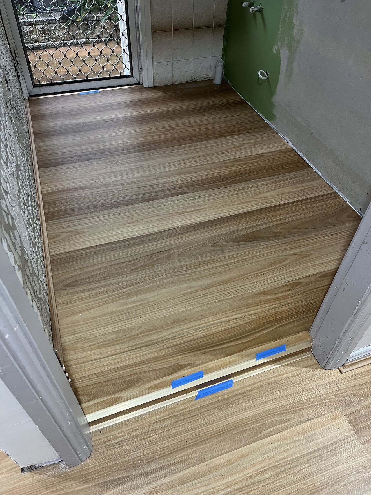 Wood flooring being installed in a doorway with a screen door in the background, and construction materials and partially painted walls visible.
