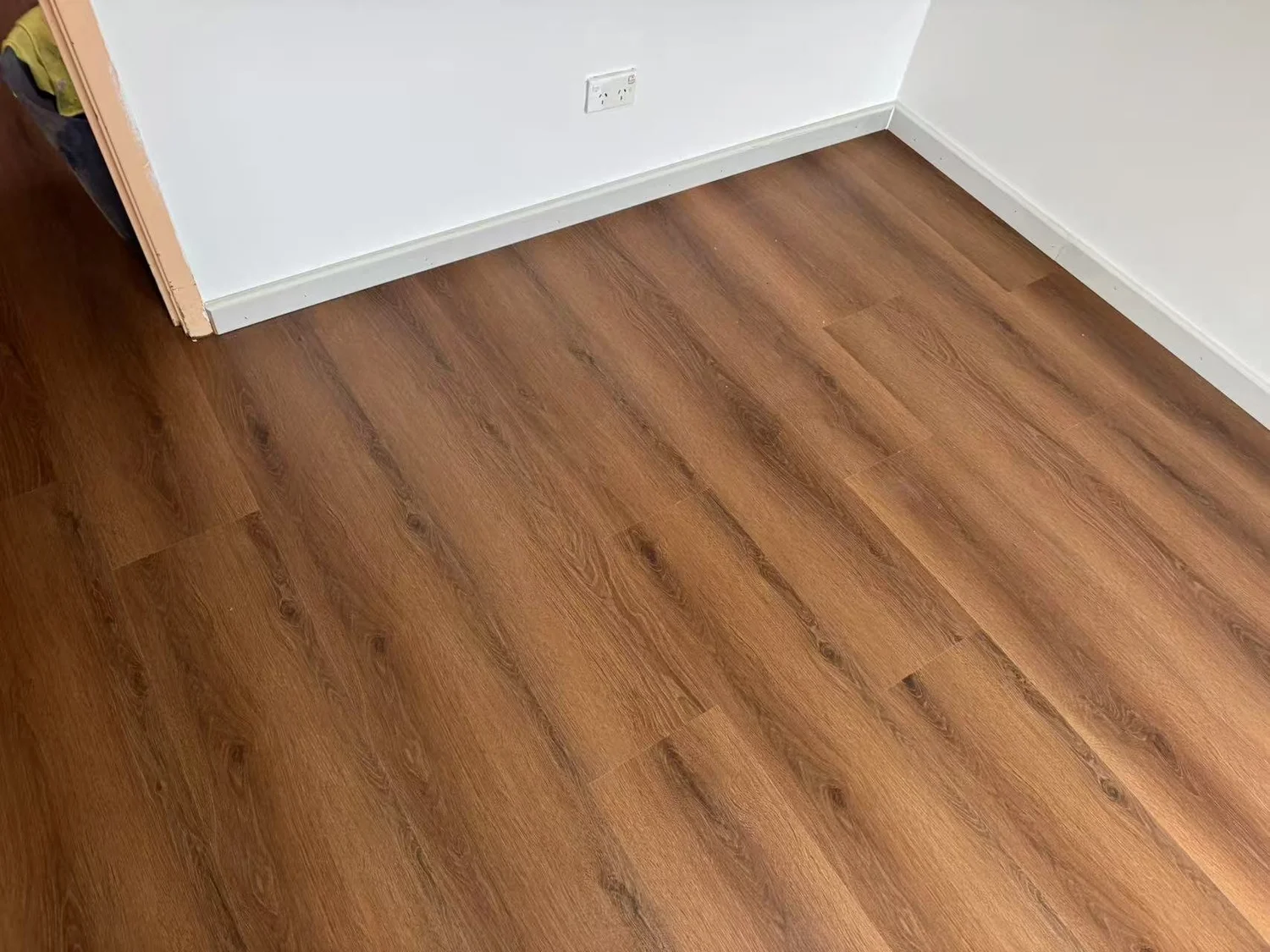 Empty room corner with wooden flooring, white walls, and a white baseboard, with a wall socket visible on the wall.