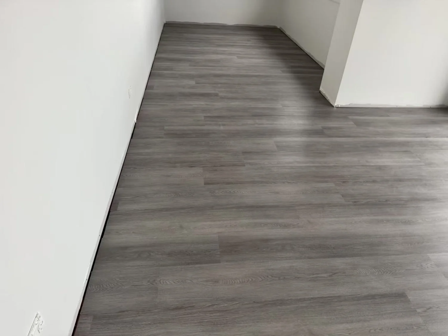 Empty room with gray wooden flooring, white walls, and a corner visible in the image.