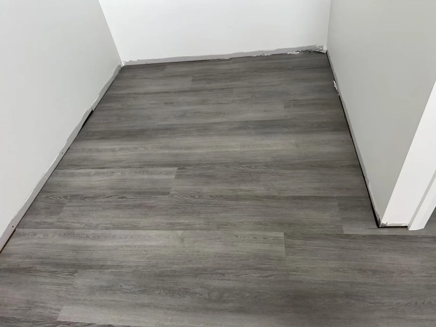 Photo of a corner of a room with light grey wood-look flooring, white walls, and baseboards, some of which appear unfinished or damaged.
