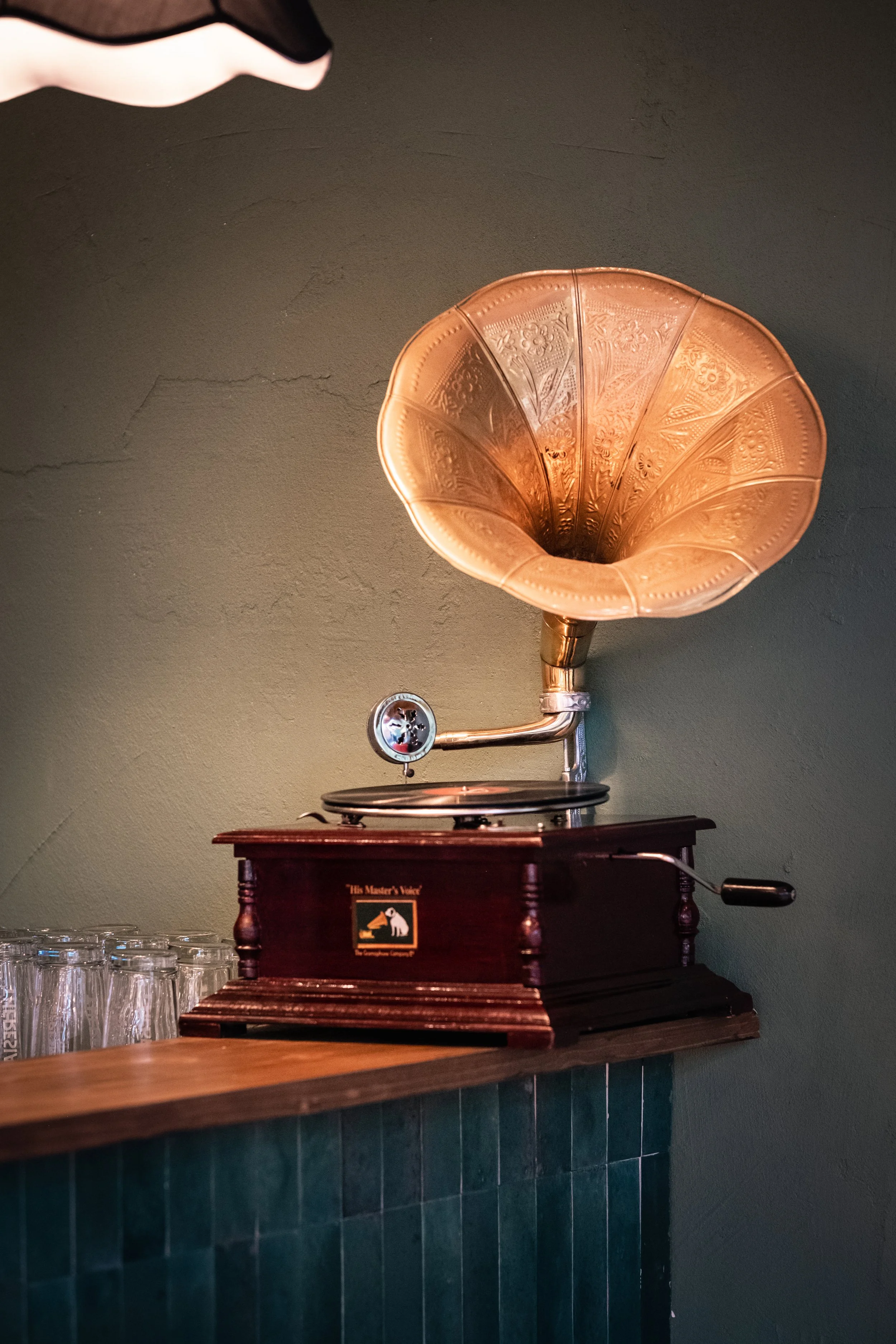 Un grammofono vintage con cassa di legno e grande coppa in metallo dorato come altoparlante, posizionato su un ripiano di legno, con bicchieri di vetro sul lato sinistro.