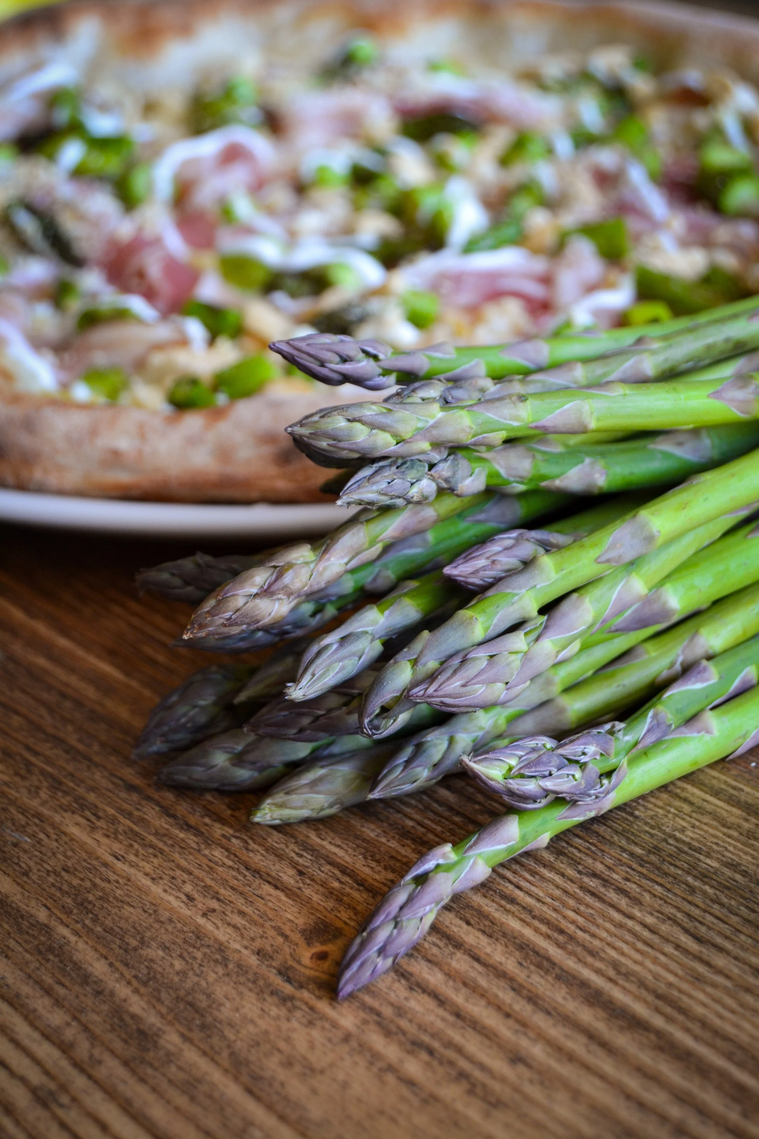 Punuevo di asparagi freschi su un tavolo di legno, con una pizza in lontananza.
