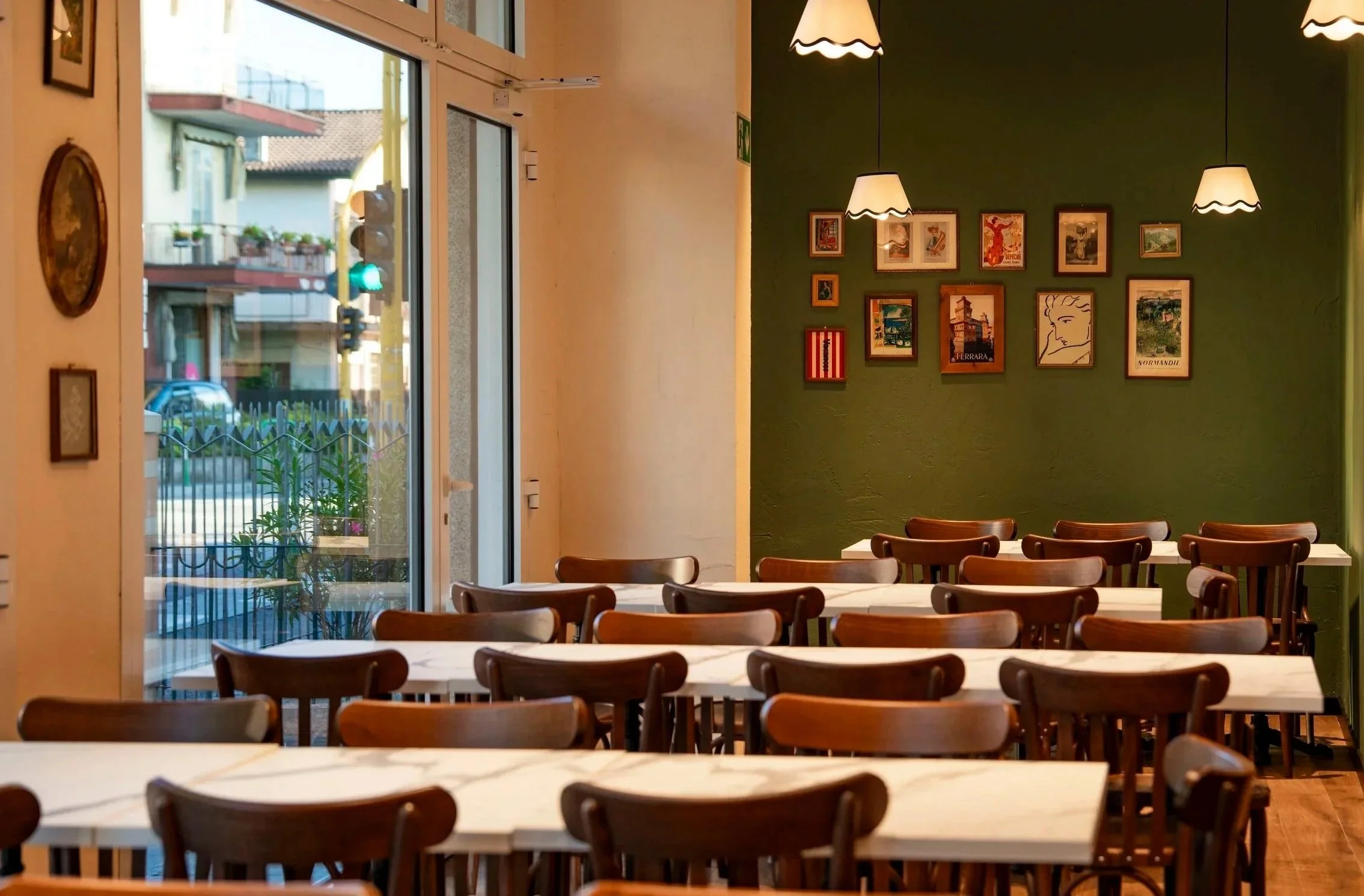 Interno di un ristorante o caffè con tavoli e sedie di legno, pareti decorate con quadri e luci sospese. Una finestra mostra la strada esterna con edifici e automobili.
