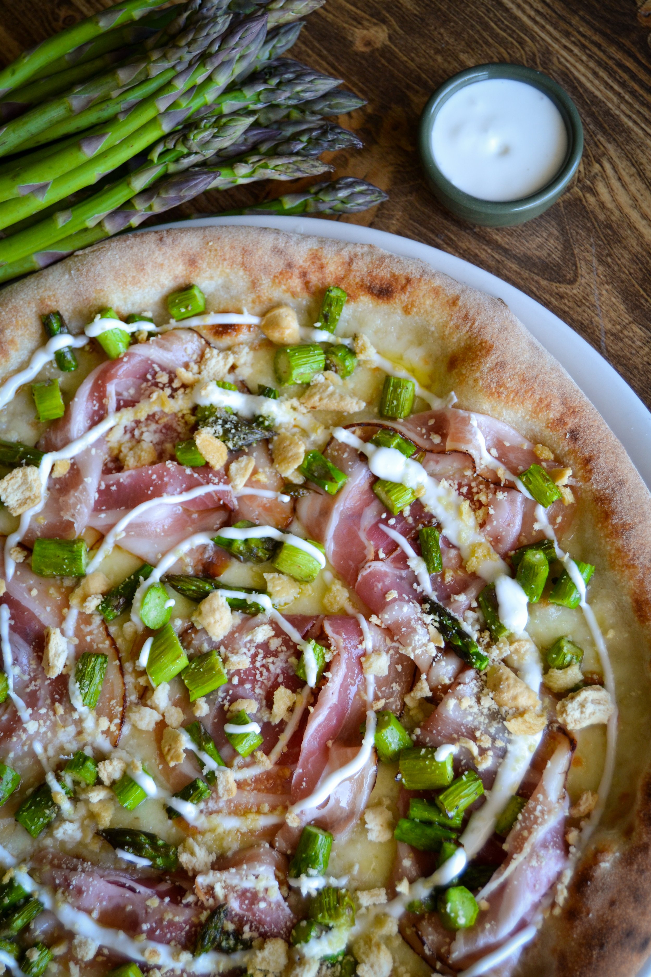 Pizza con prosciutto, asparagi, salsa bianca, e pangrattato su una teglia. Accanto, asparagi freschi e una ciotola di salsa bianca.