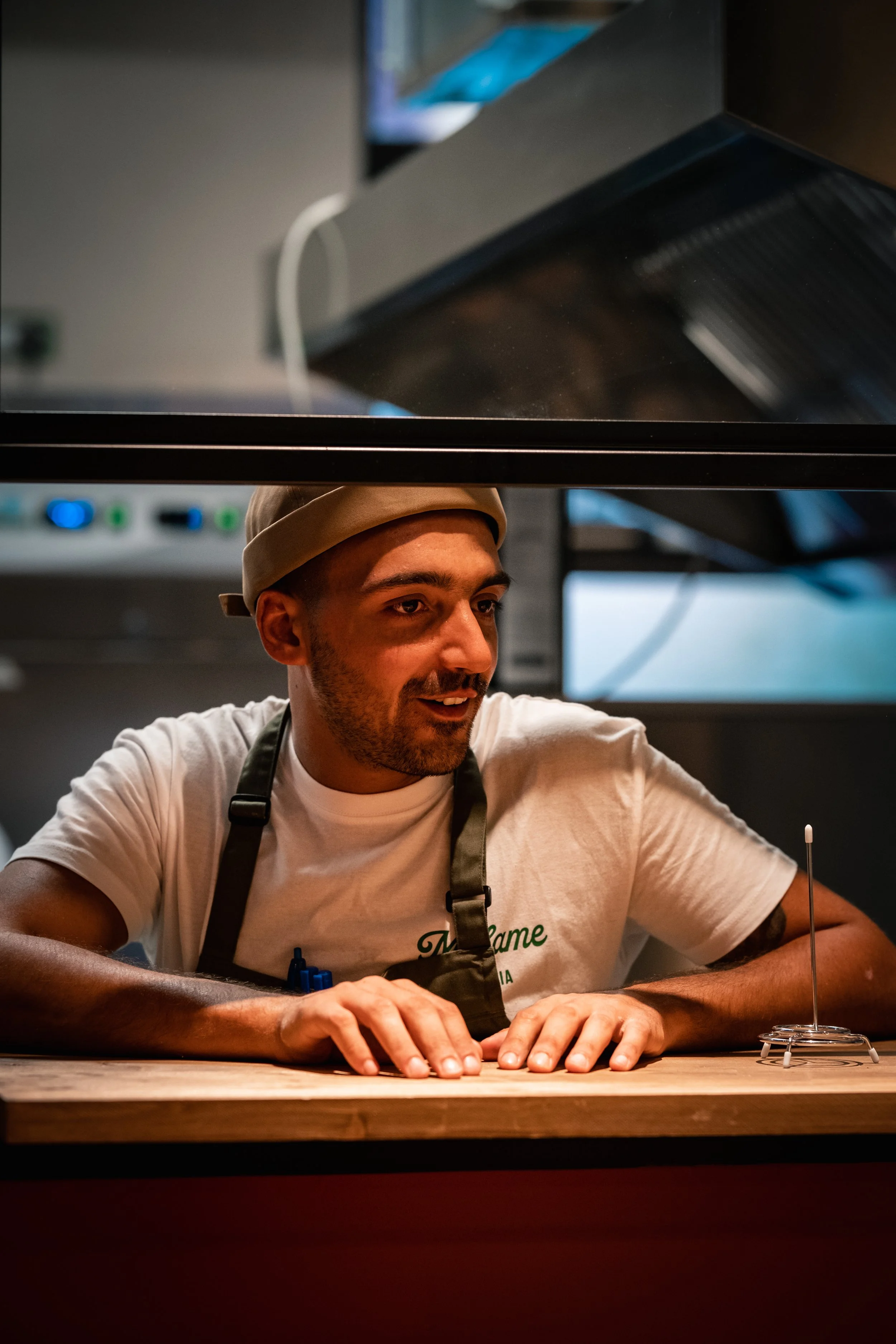 Un uomo sorridente, con cappello e grembiule, guarda attraverso una finestra in una cucina o laboratorio, con attrezzature professionali sullo sfondo.