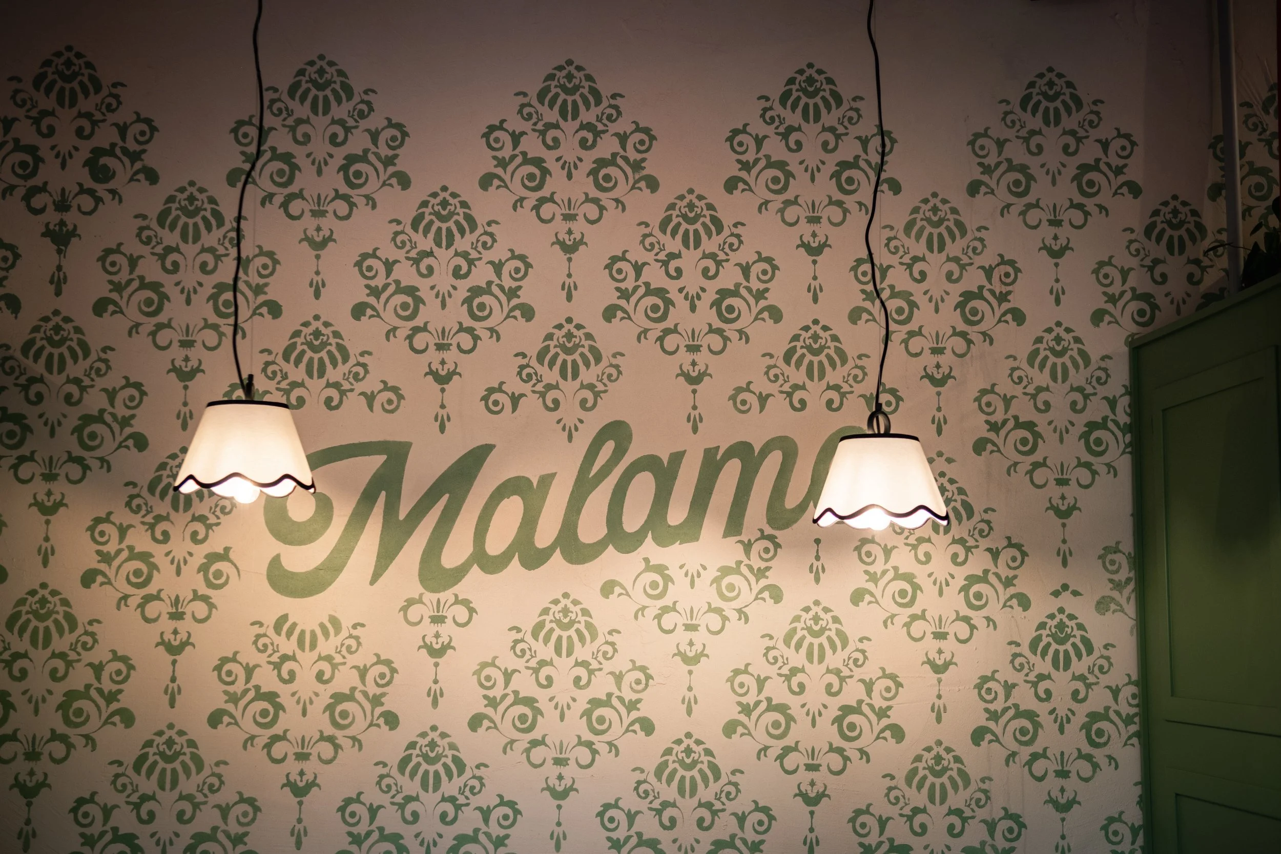 Mural con il nome 'Malami' scritto su una parete decorata con un motivo floreale verde e due lampade accese appese al soffitto.