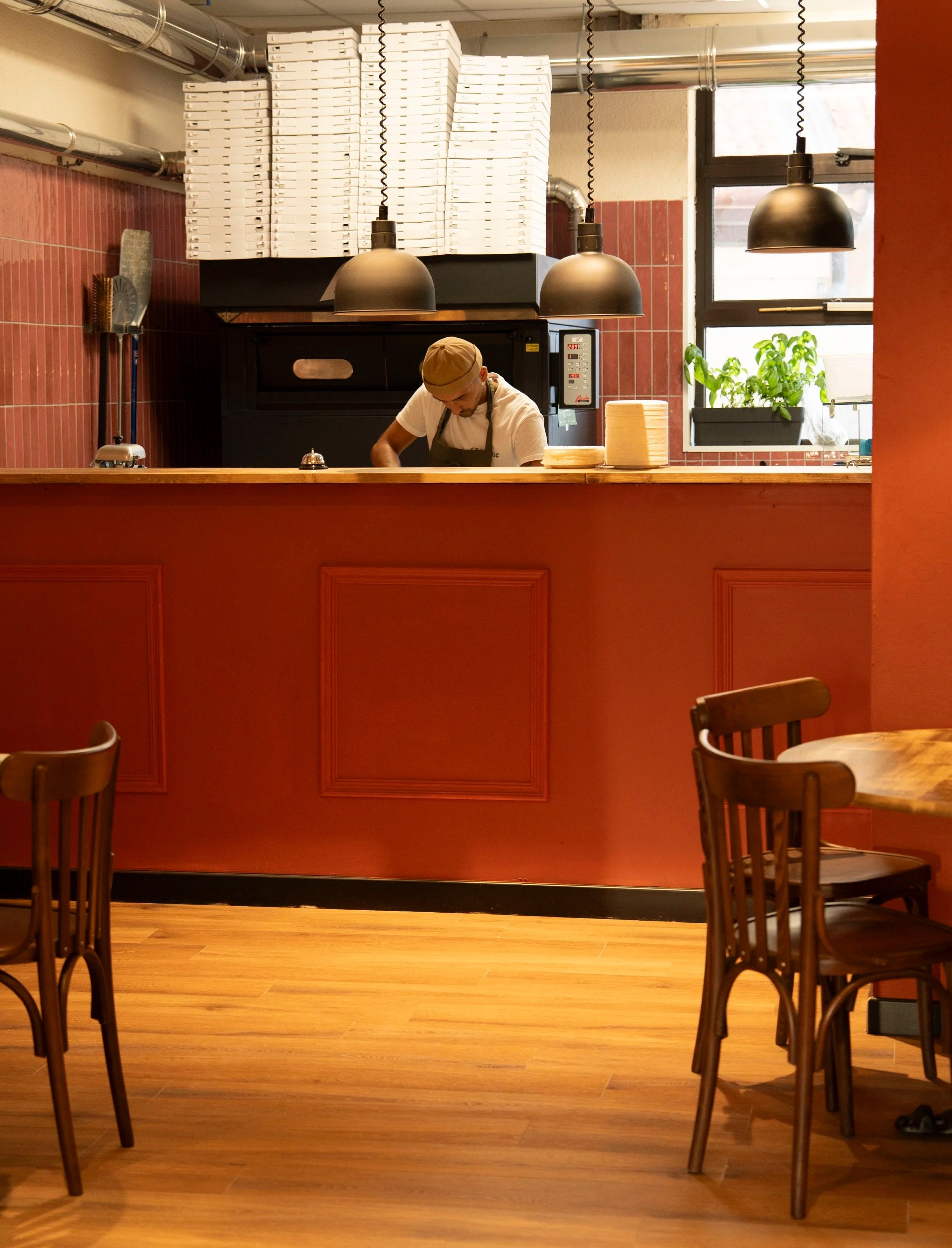 Interno di una pizzeria o ristorante con un cuoco che lavora dietro il bancone, scaffali con scatole di pizza surgelata, lampade nere, pareti rosse, tavoli e sedie di legno, finestra con pianta sul davanzale.