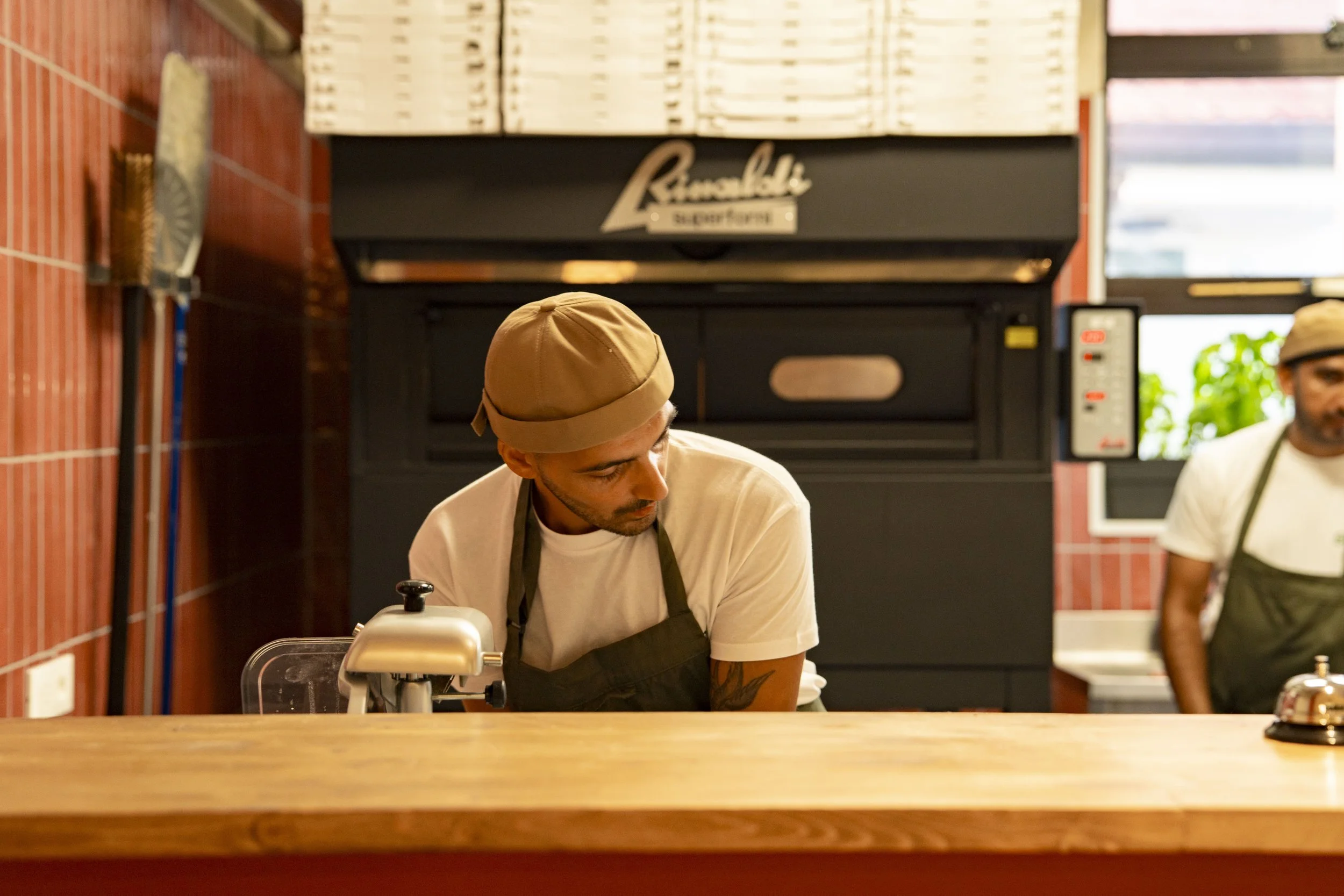 Un barista con berretto e grembiule nero si inclina mentre lavora dietro un bancone in un caffè.