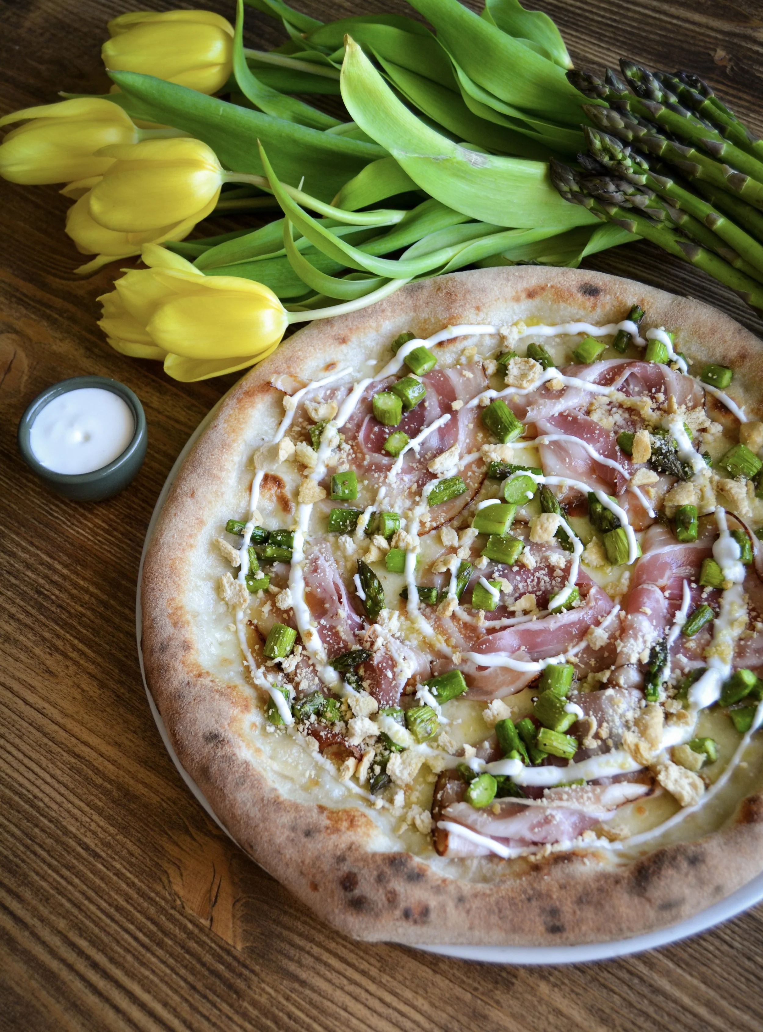 Pizza con prosciutto, funghi e cipolle verdi, con contorno di tulipani gialli e asparagi verdi, su tavola di legno.
