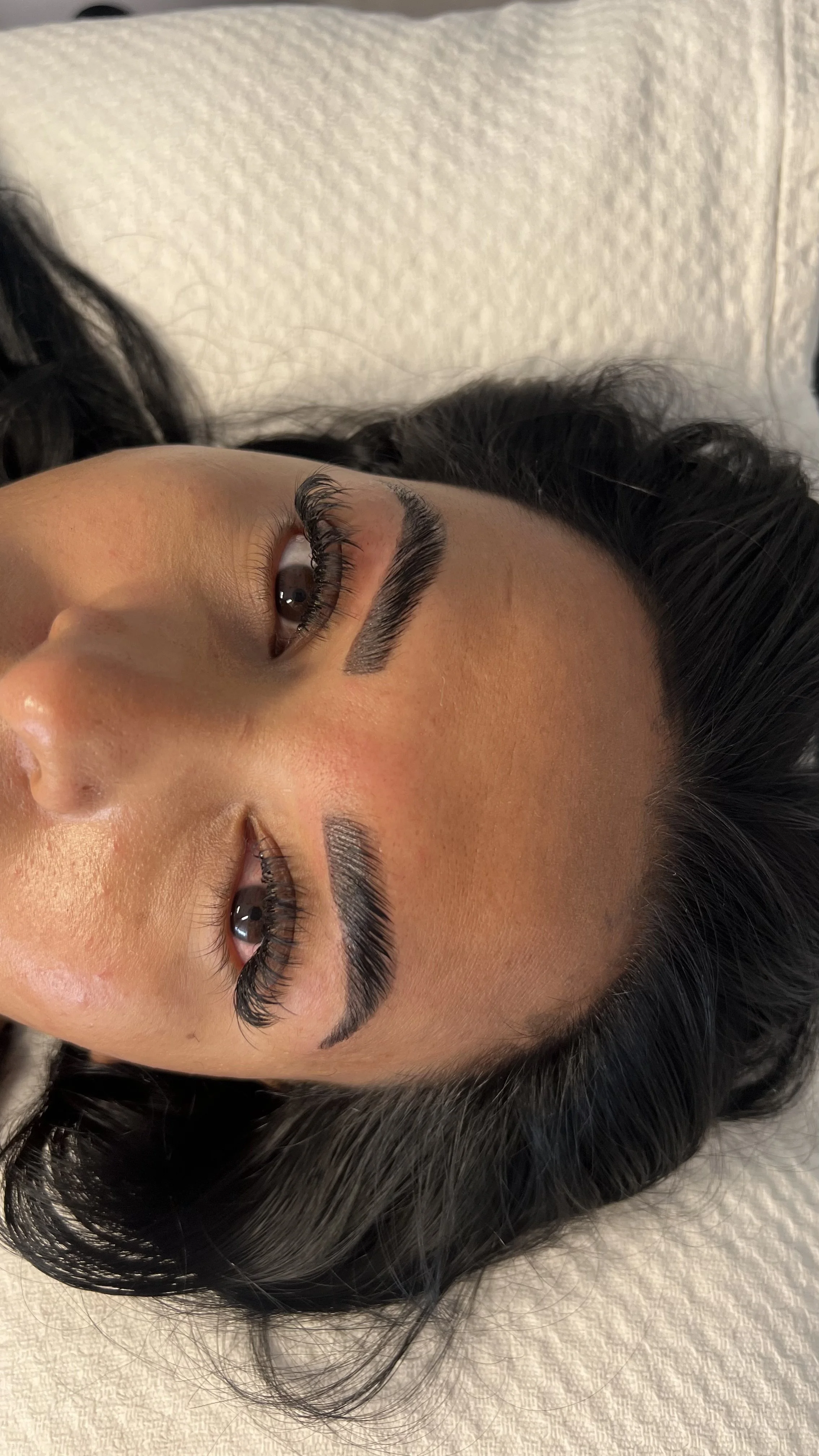 A woman with long dark hair lying on a white textured surface, showcasing her well-groomed eyebrows and long eyelashes.