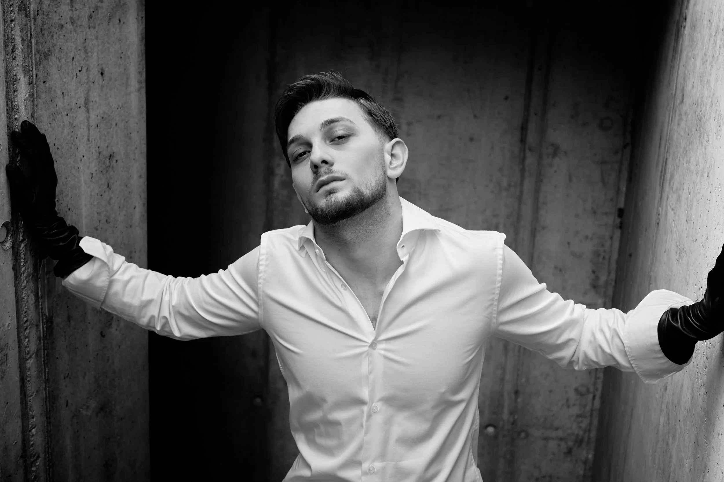 Schwarz-weiße Fotografie eines jungen Mannes mit dunklem Haar, der in einem weißen Hemd und schwarzen Handschuhen an einer rauen Betonwand steht.