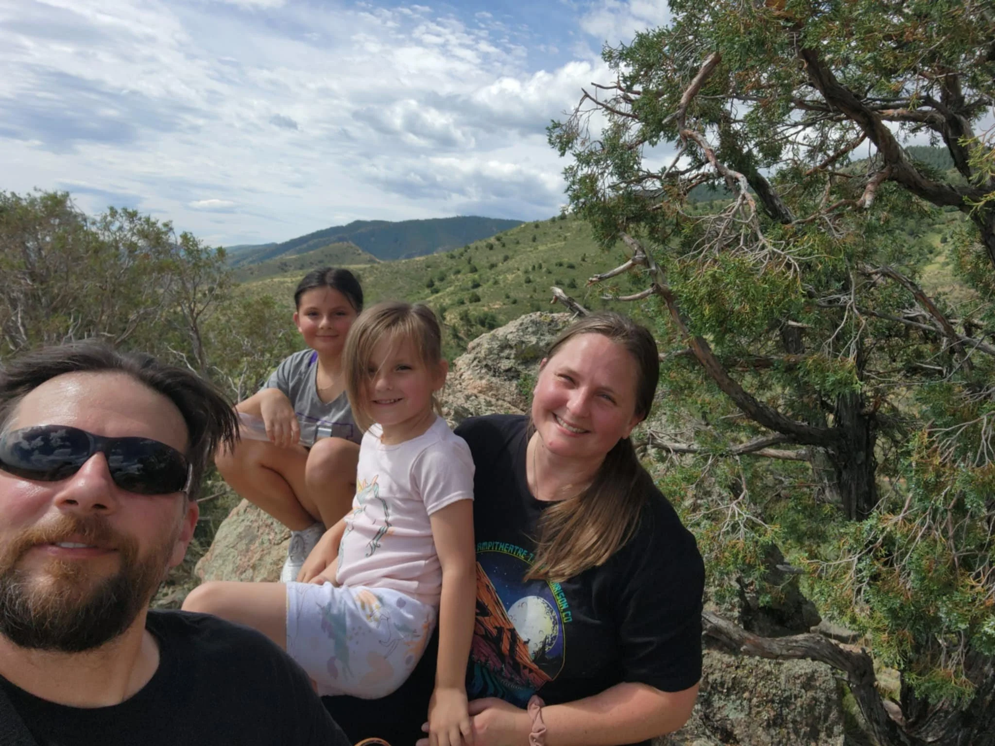 A group of four people, consisting of an adult man, two children, and an adult woman, smiling and sitting on rocks in a mountainous landscape. Trees and a clear blue sky with clouds are in the background.