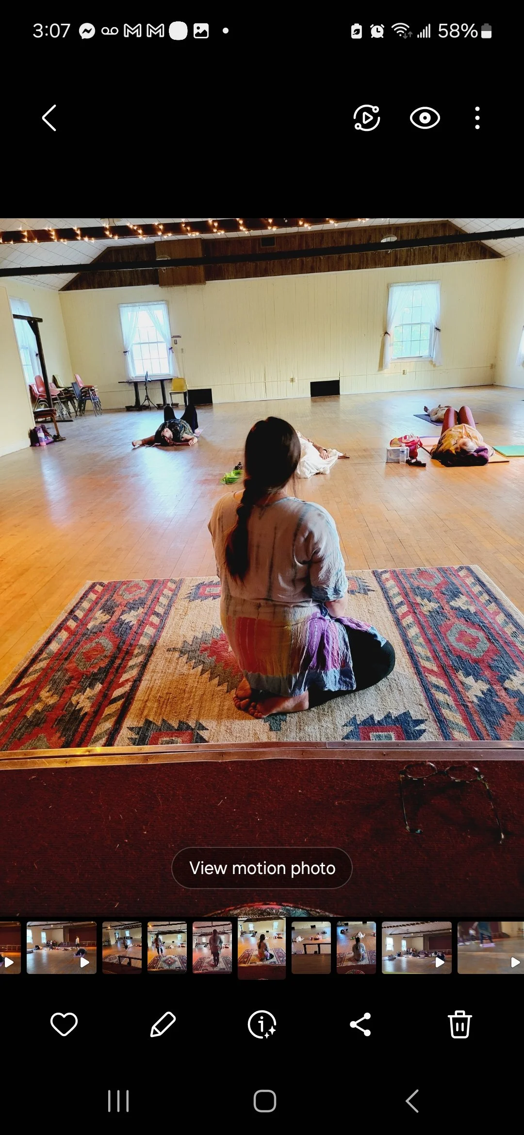Indoor room with people practicing breathwork meditation, one sitting on a rug, others lying on mats, with a wooden floor and a few windows with drapes.