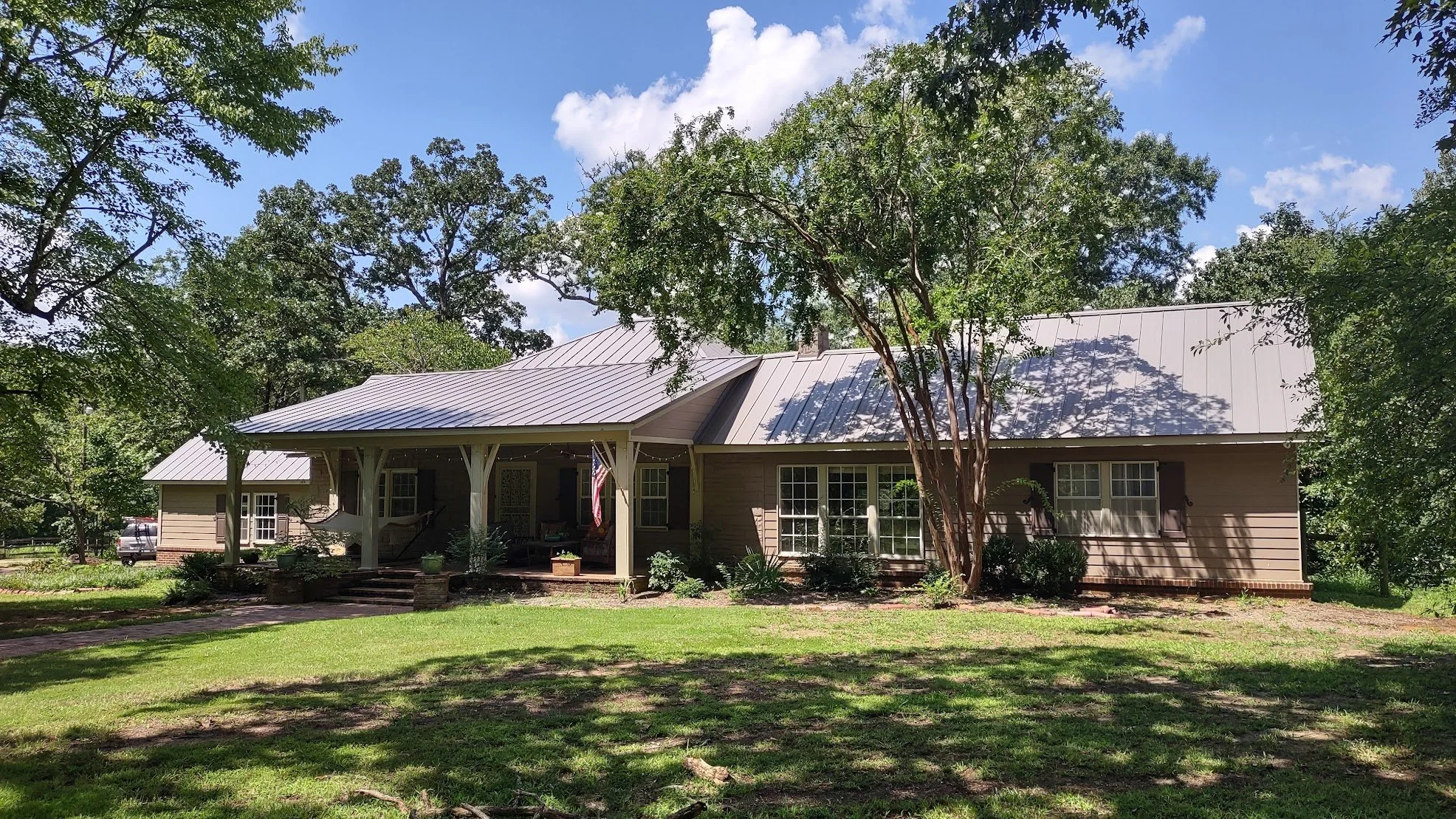 Single-story house with a metal roof and porch, surrounded by trees and grass, under a blue sky with clouds.