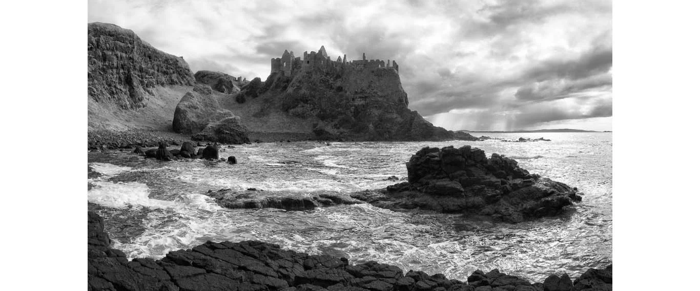 Black and white photograph of the ruins of Dunluce Castle on the rocky coastline of Northern Ireland, with dramatic skies.