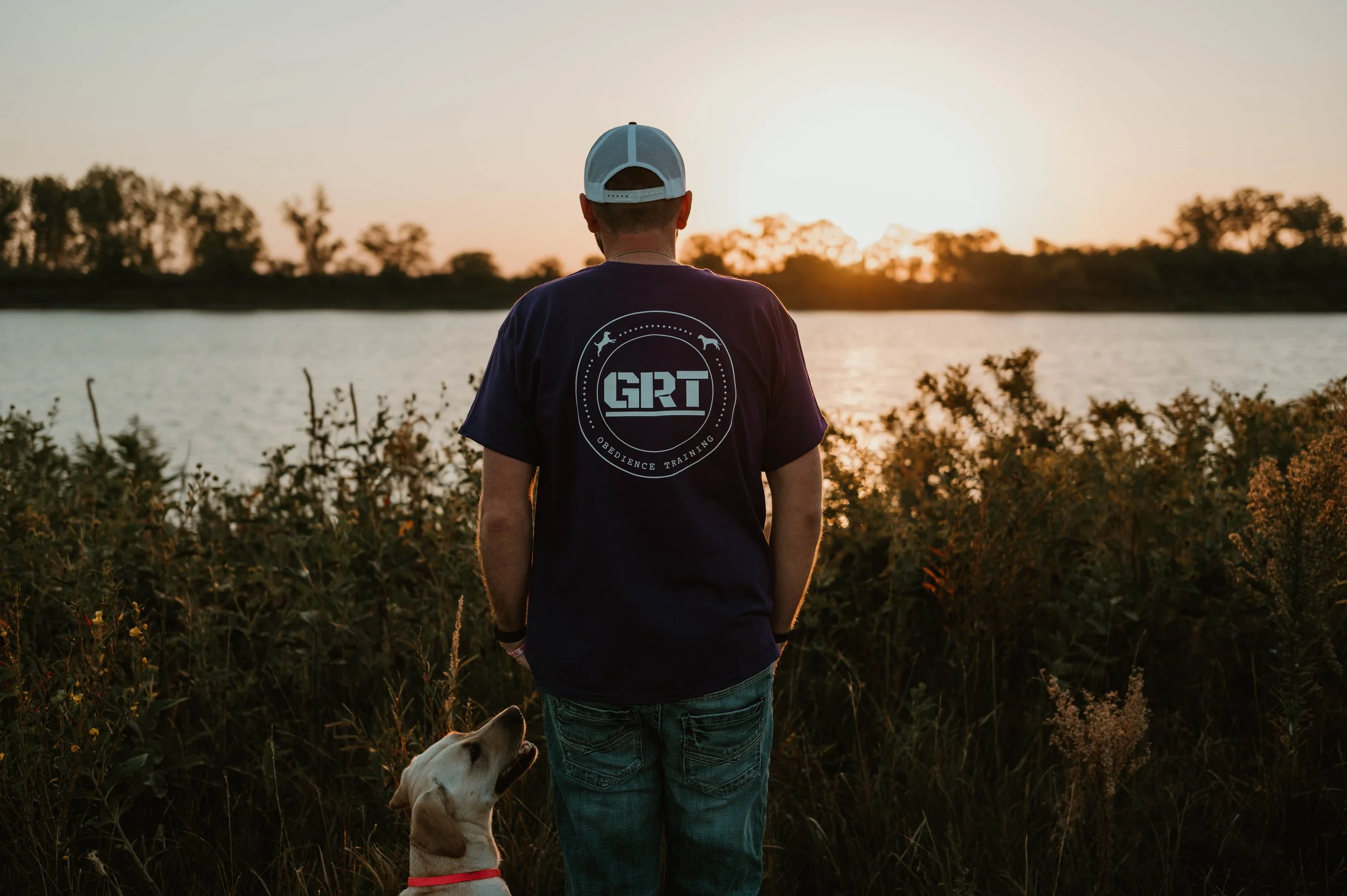 A man standing near a lake at sunset, wearing a navy T-shirt with 'GRT' and 'Obedience Training' printed on the back, with a yellow lab retriever sitting beside him and looking up at him.