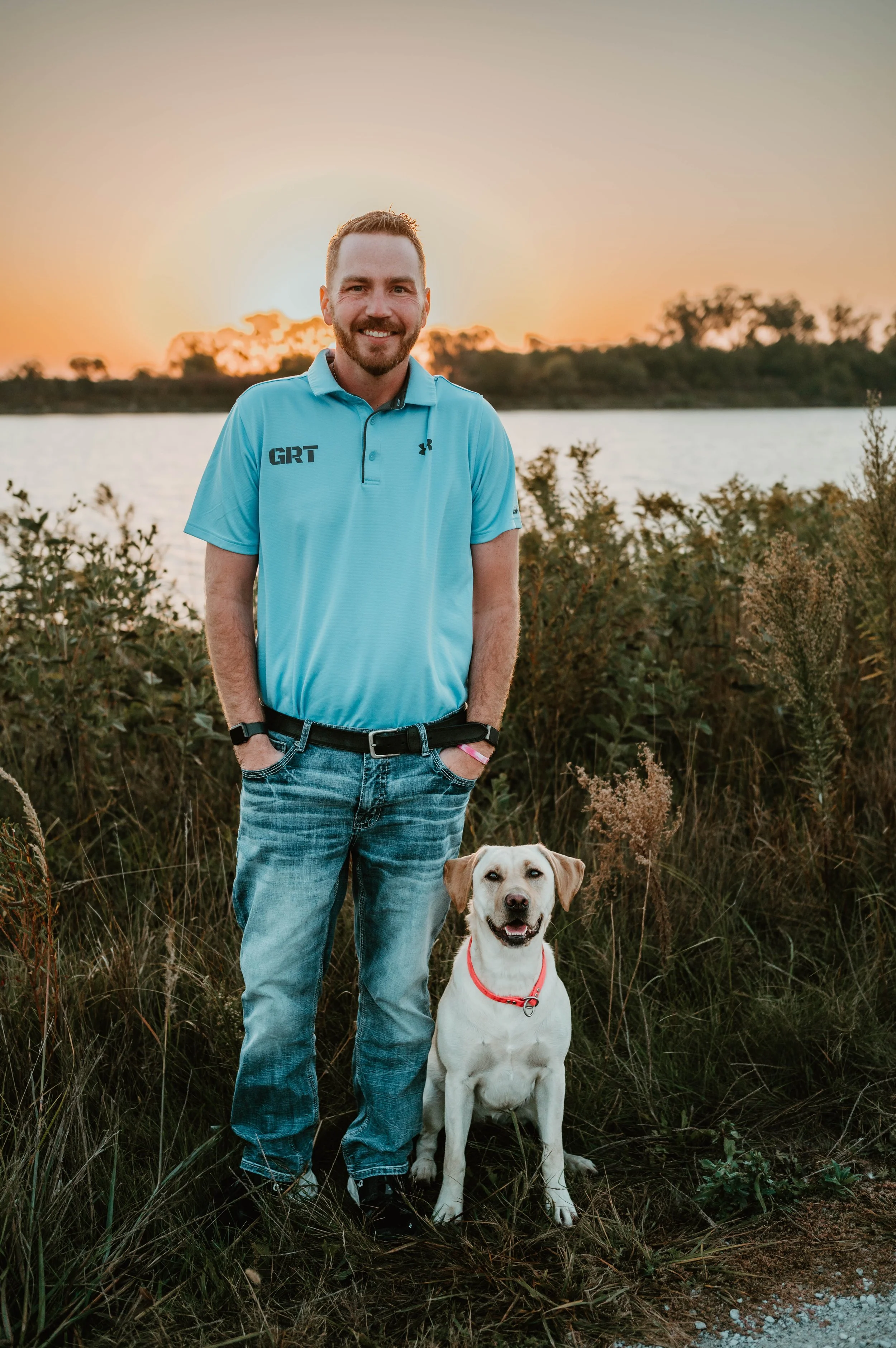 A man in a blue polo shirt and jeans standing outdoors with a dog by a body of water during sunset.