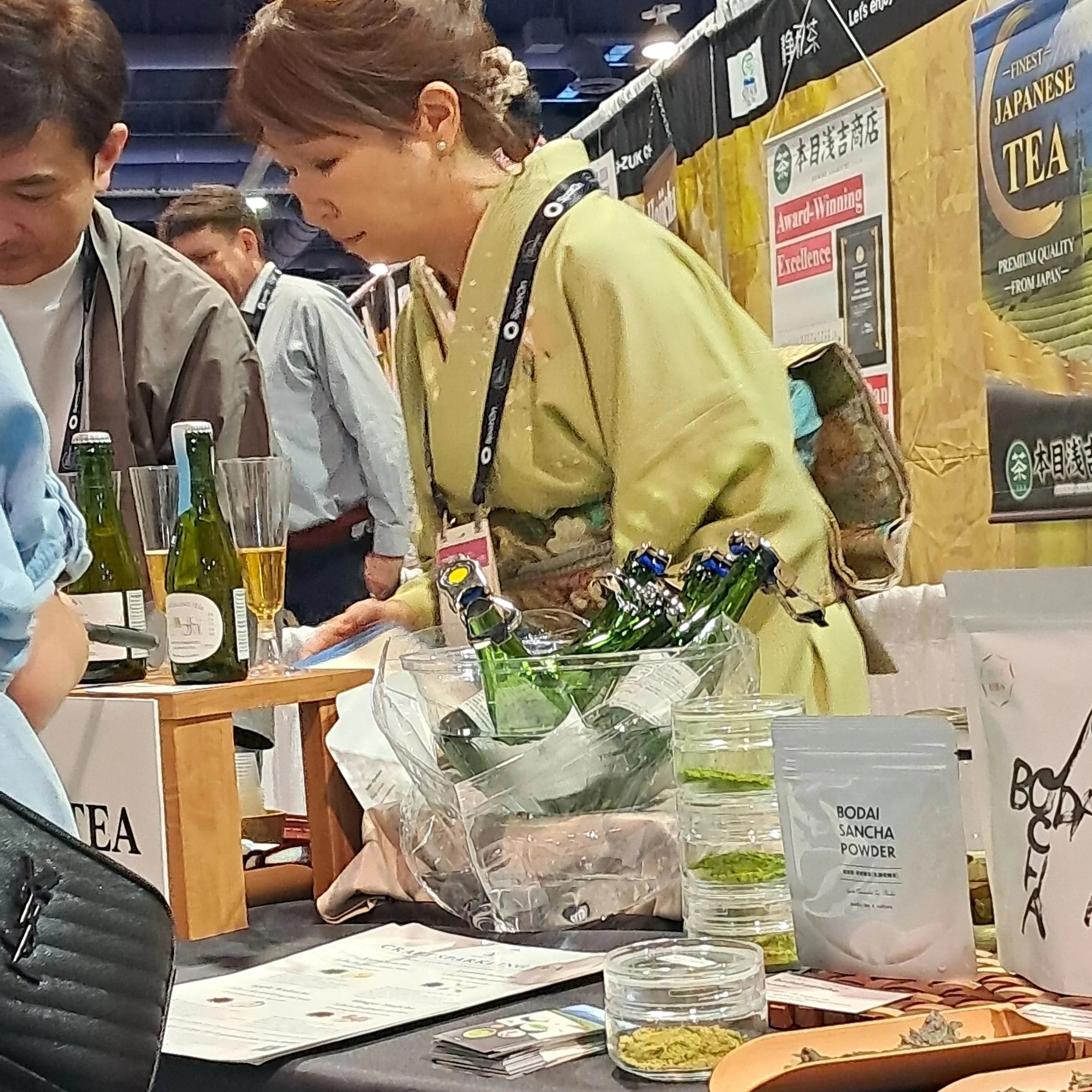 A table arranged with loose leaf tea displays, packaged teas, and glass bottles of sparkling teas, behind which stands a woman dressed in green kimono and intricate brocade obi.