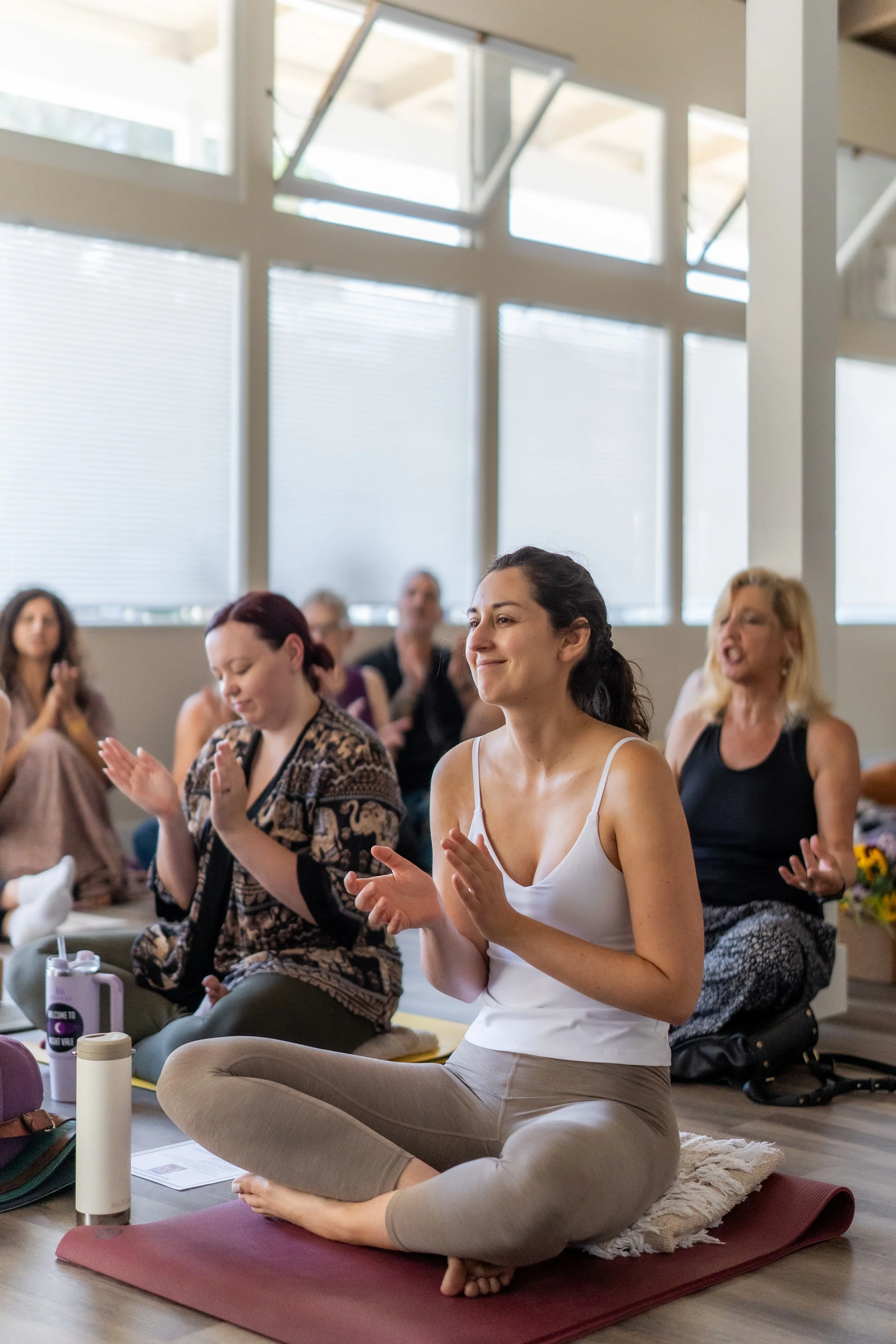 Group of women participating in a meditation or yoga class indoors, sitting cross-legged on mats with eyes closed or relaxed, some clapping, with large windows allowing natural light to fill the room.